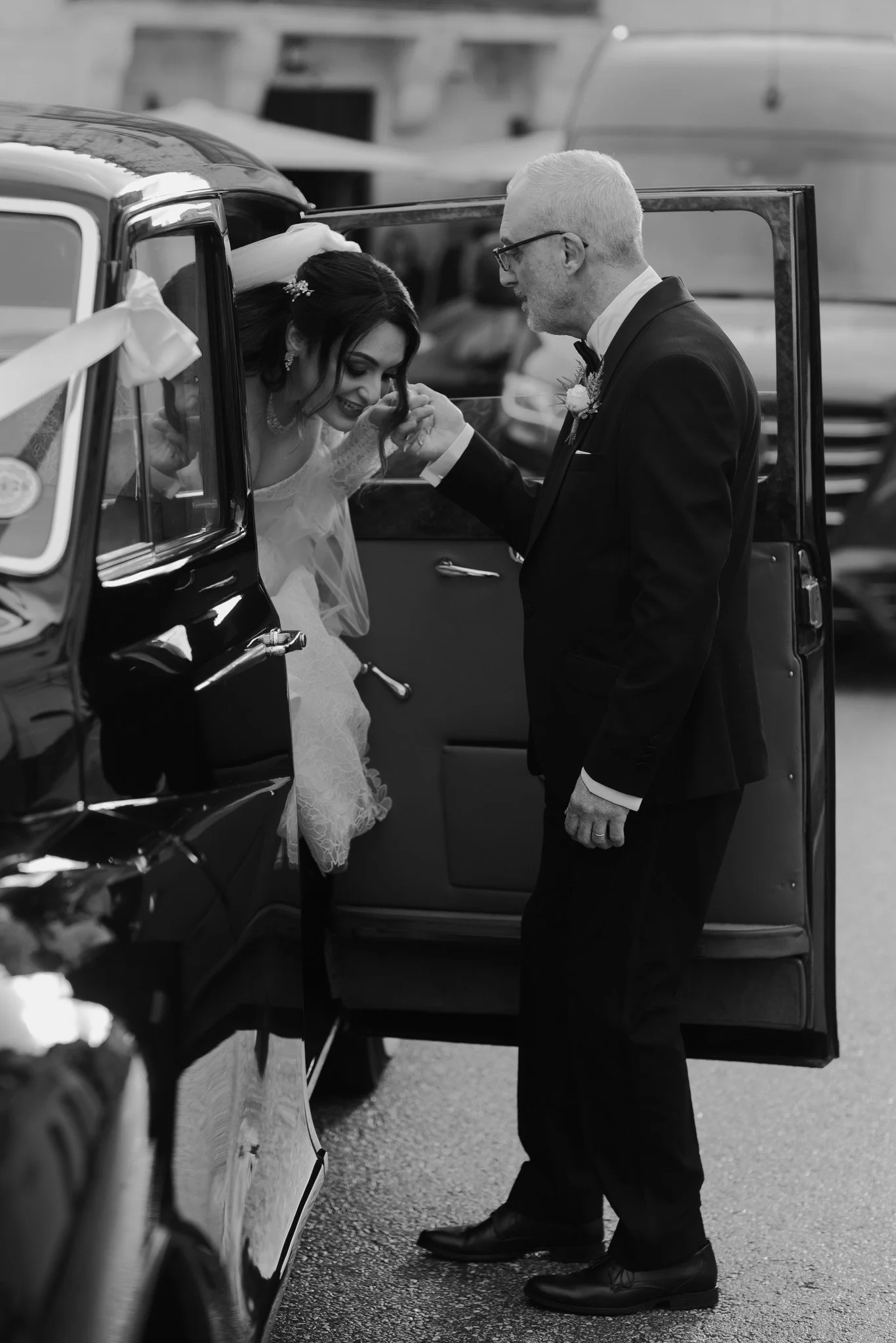 A groom in a tuxedo helping a bride in a wedding dress out of a vintage car.
