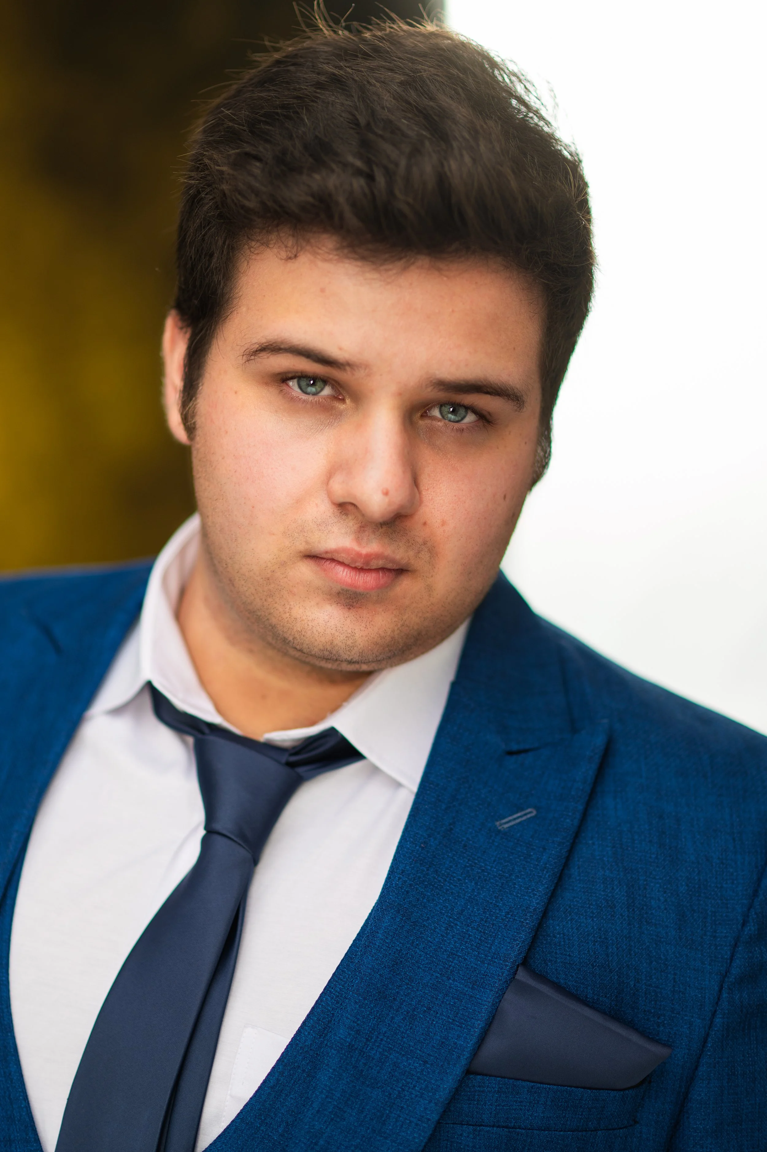 Close-up of a young man with dark hair and blue eyes wearing a blue suit jacket, white shirt, and dark tie, with a white background.