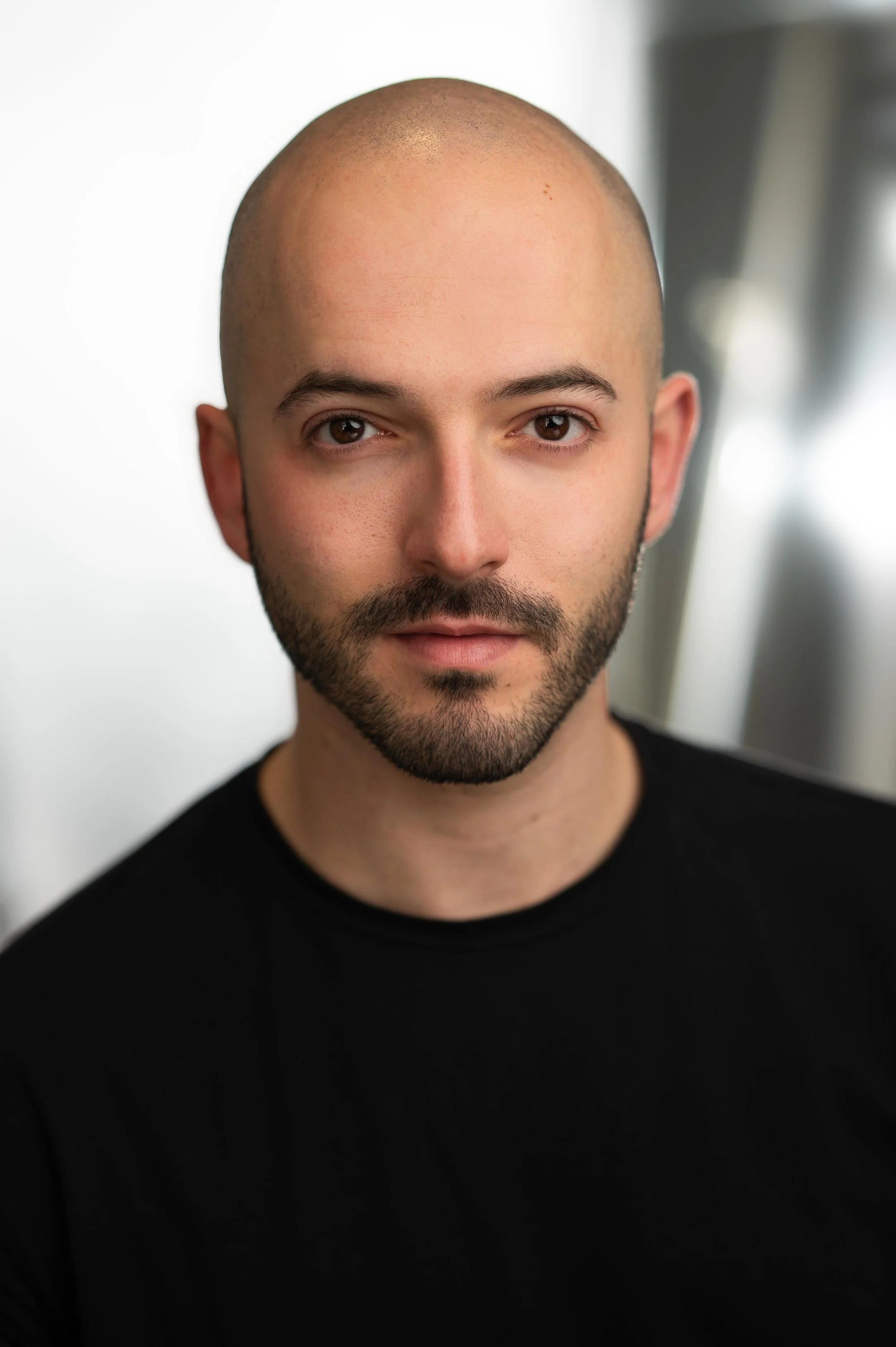 Close-up portrait of a bald man with a beard, wearing a black shirt, looking directly at the camera with a neutral expression, background slightly blurred.
