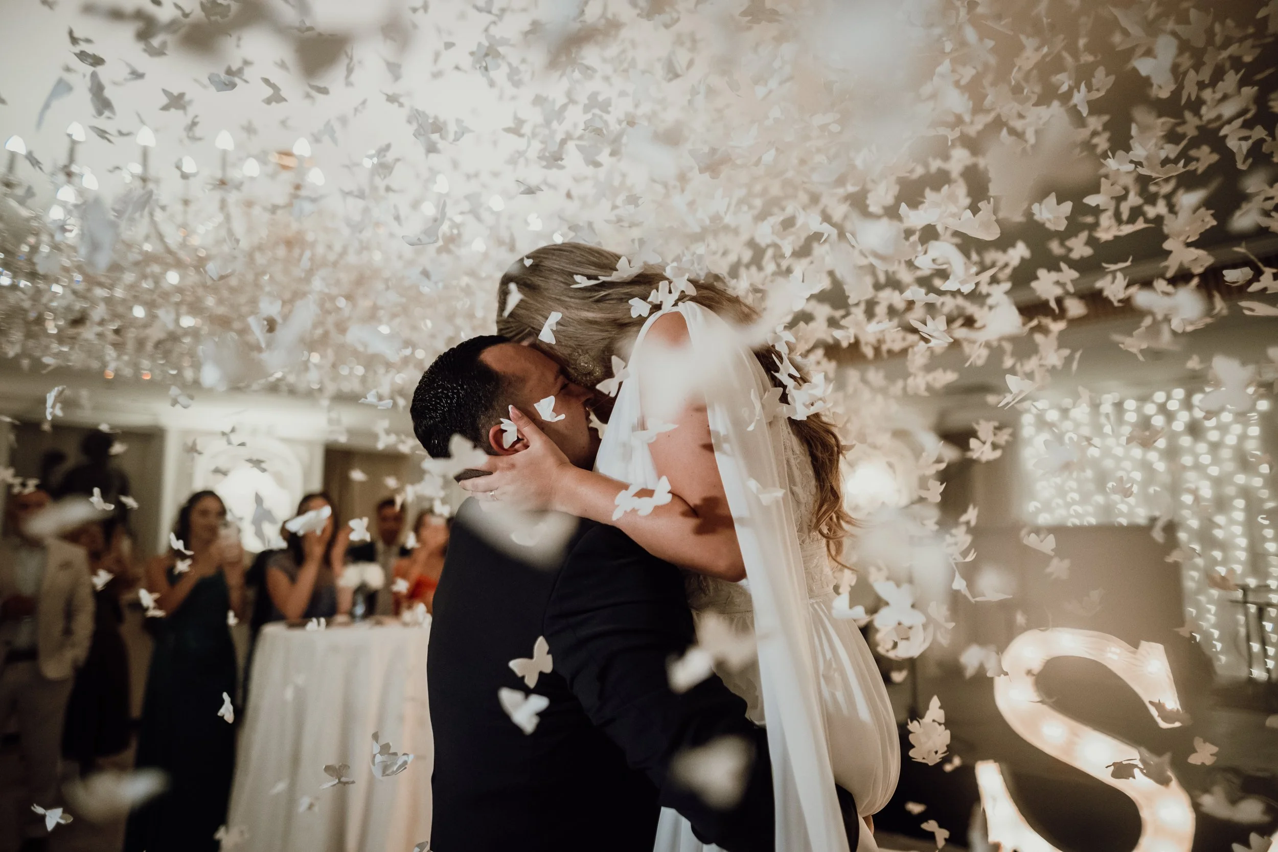 A bride and groom kiss during their wedding reception, surrounded by falling white paper butterflies and a large chandelier overhead, with guests in the background watching.
