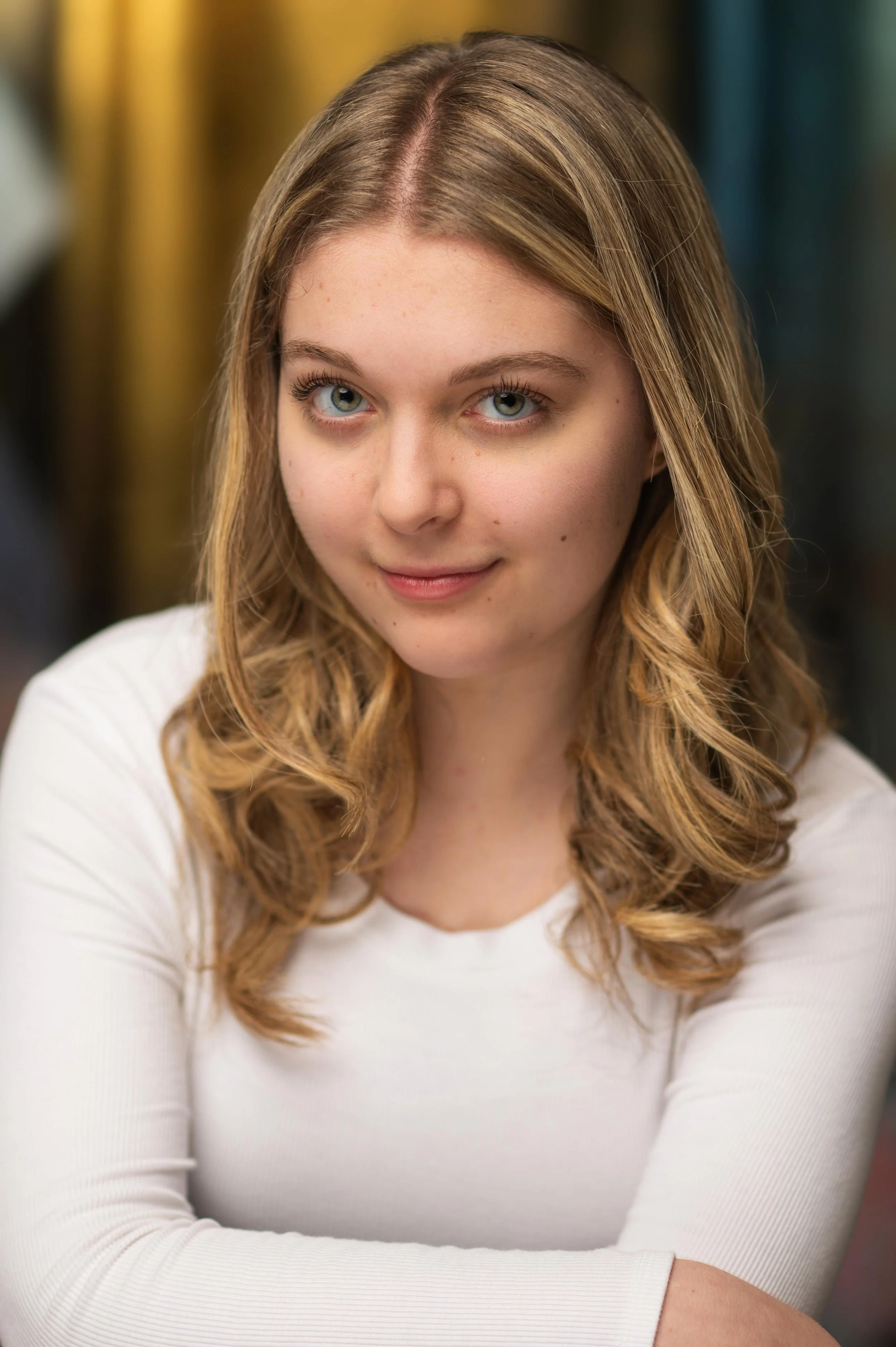 Close-up of a young woman with light brown wavy hair, blue eyes, wearing a white long-sleeve shirt, smiling subtly, with a blurred background.
