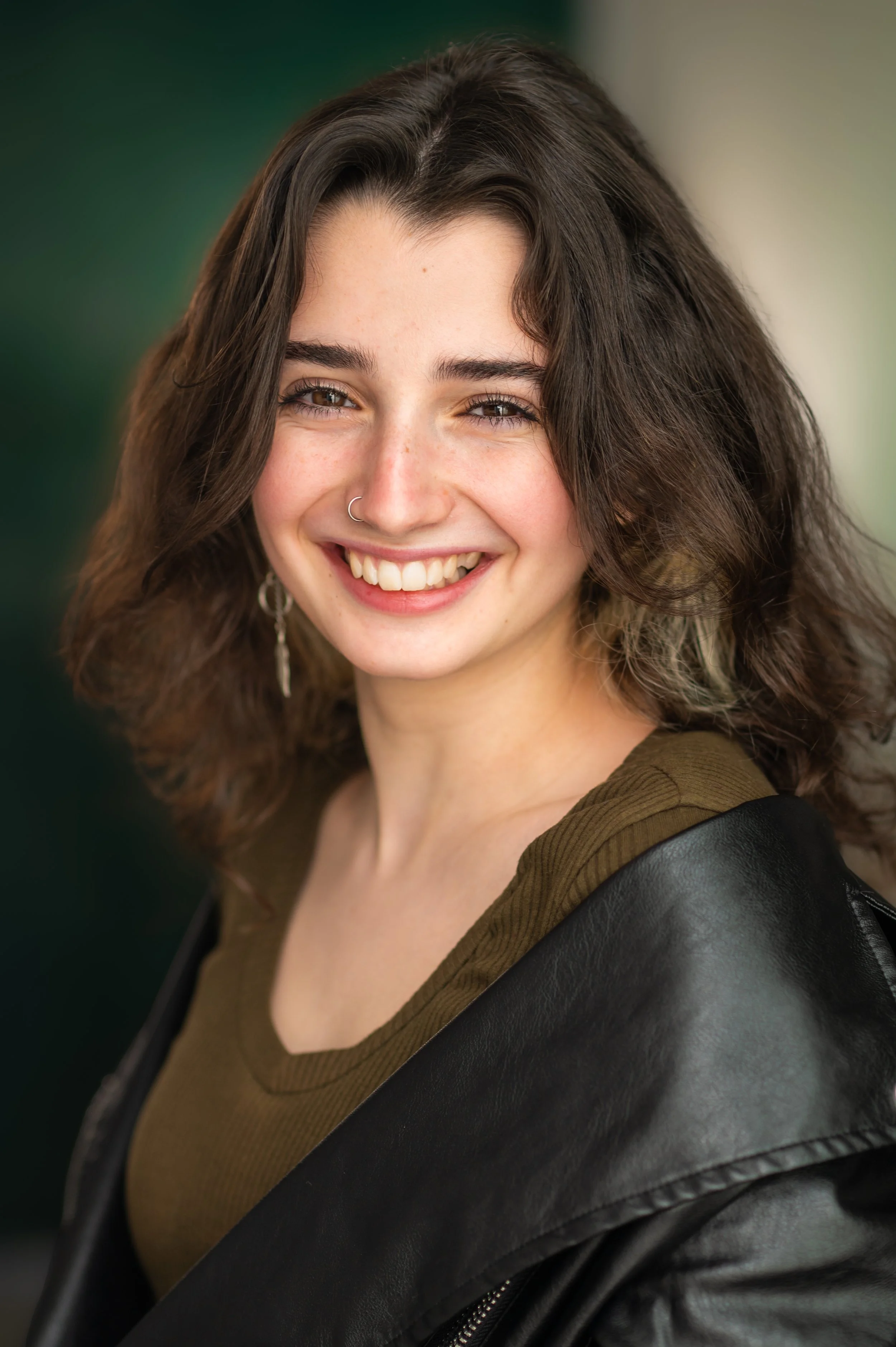 Close-up of a young woman with long brown hair, smiling, with a nose ring, wearing a brown top and a black leather jacket, against a blurred green background.