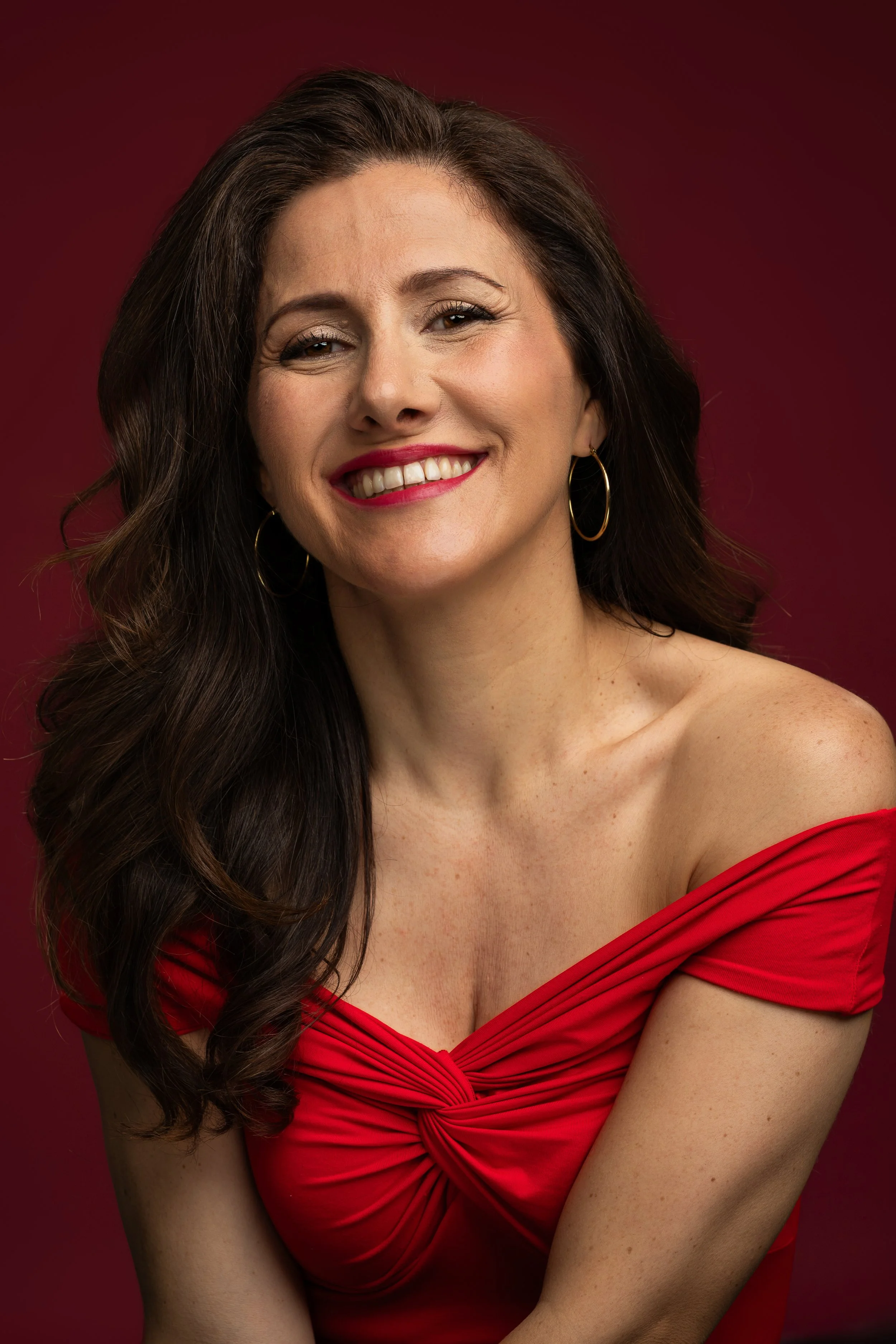 A woman with long, dark, wavy hair smiling, wearing red off-the-shoulder dress and gold hoop earrings, against a solid dark red background.