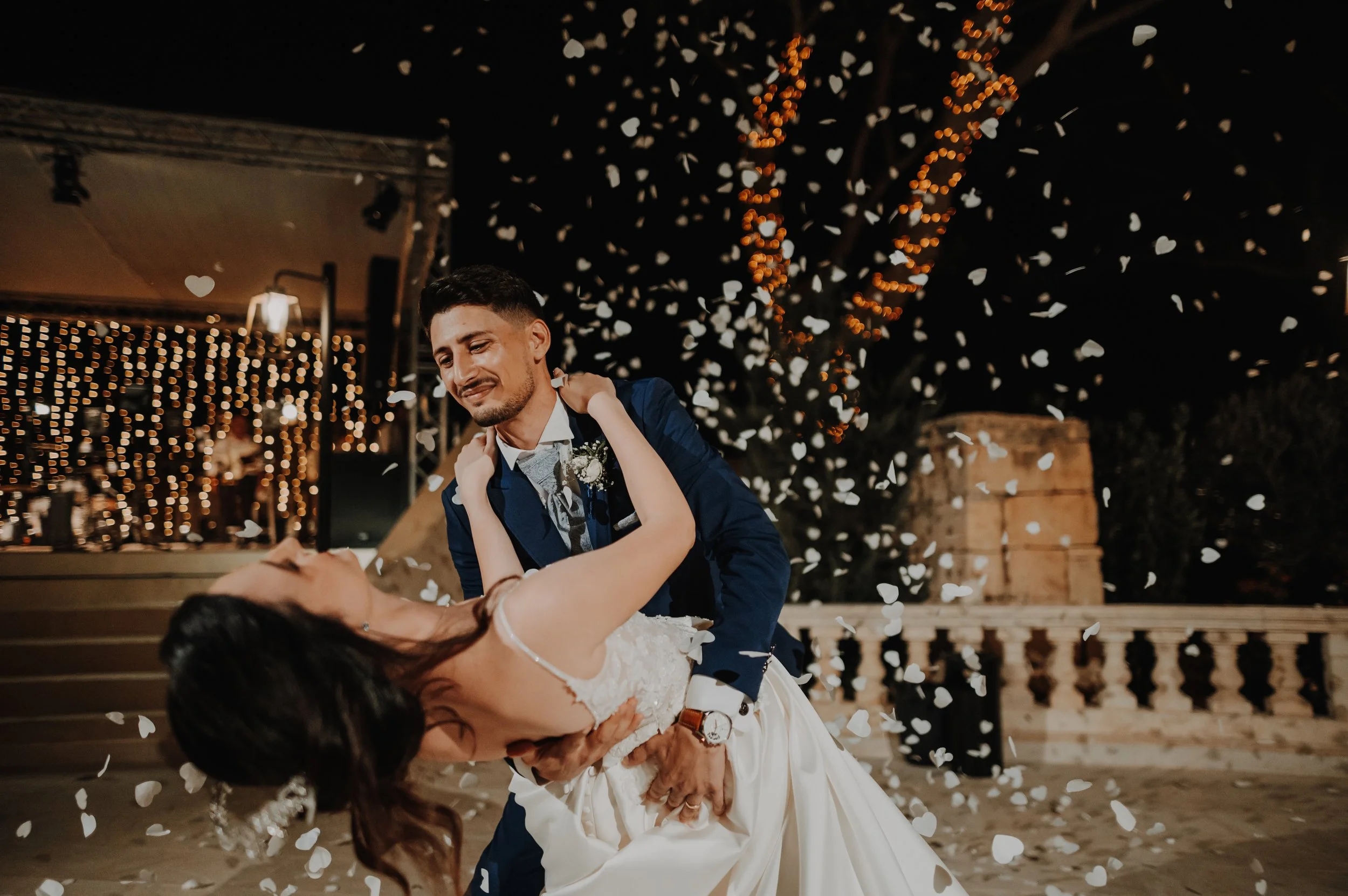 A groom in a blue suit dancing with a bride in a white wedding dress surrounded by falling flower petals at night with string lights in the background.