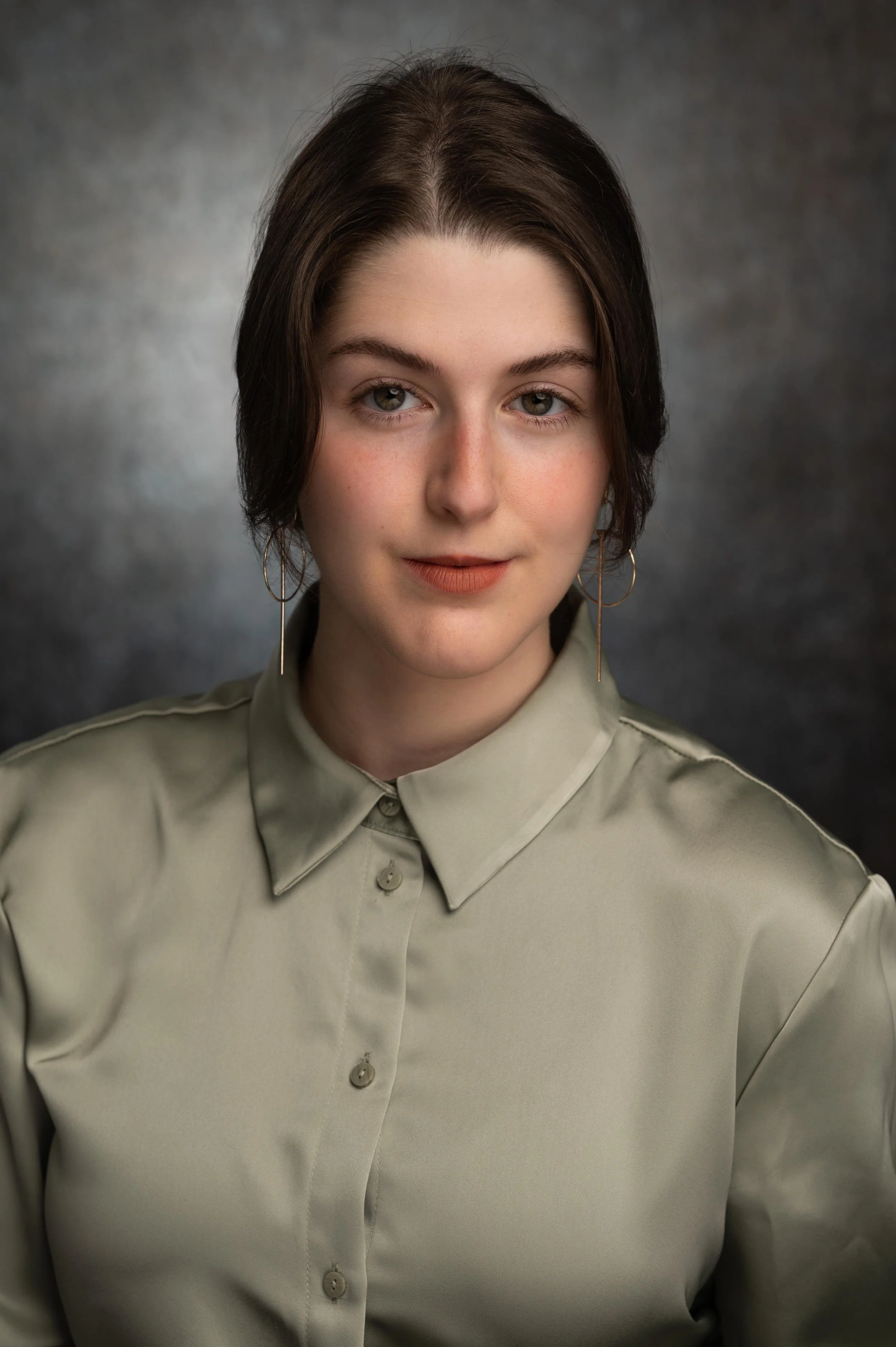 Close-up portrait of a young woman with short dark brown hair and hazel eyes, wearing gold hoop earrings and a khaki button-up shirt, against a dark blurred background.