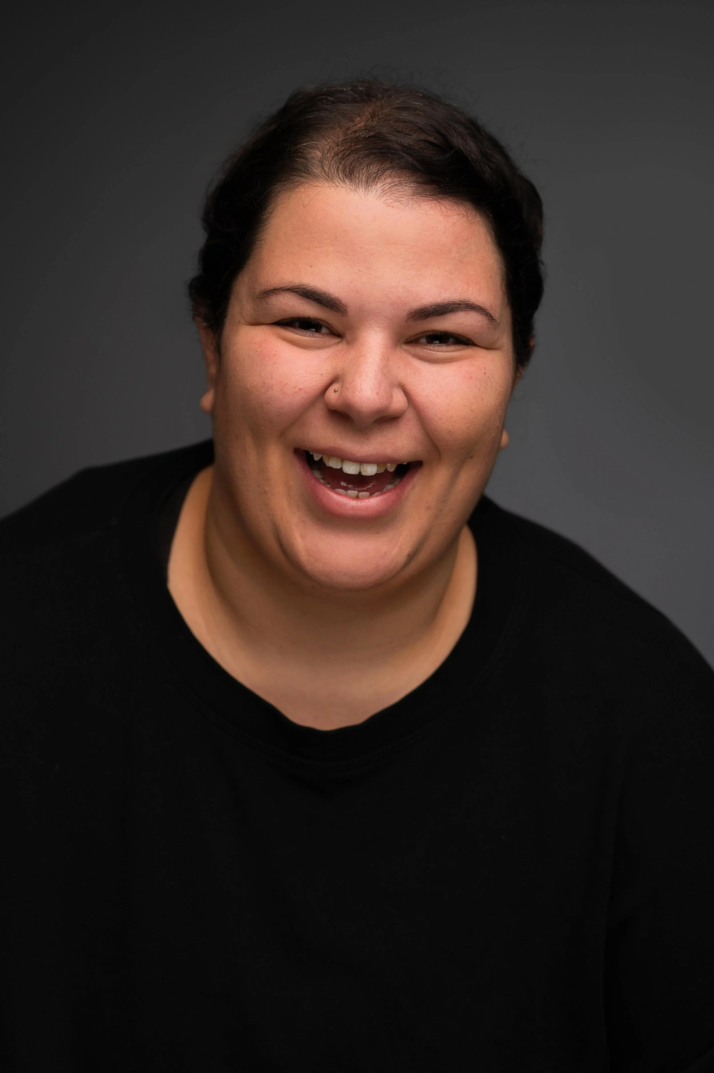 Close-up of a smiling woman with short dark curly hair, wearing a black top, and a small nose piercing, against a dark grey background.