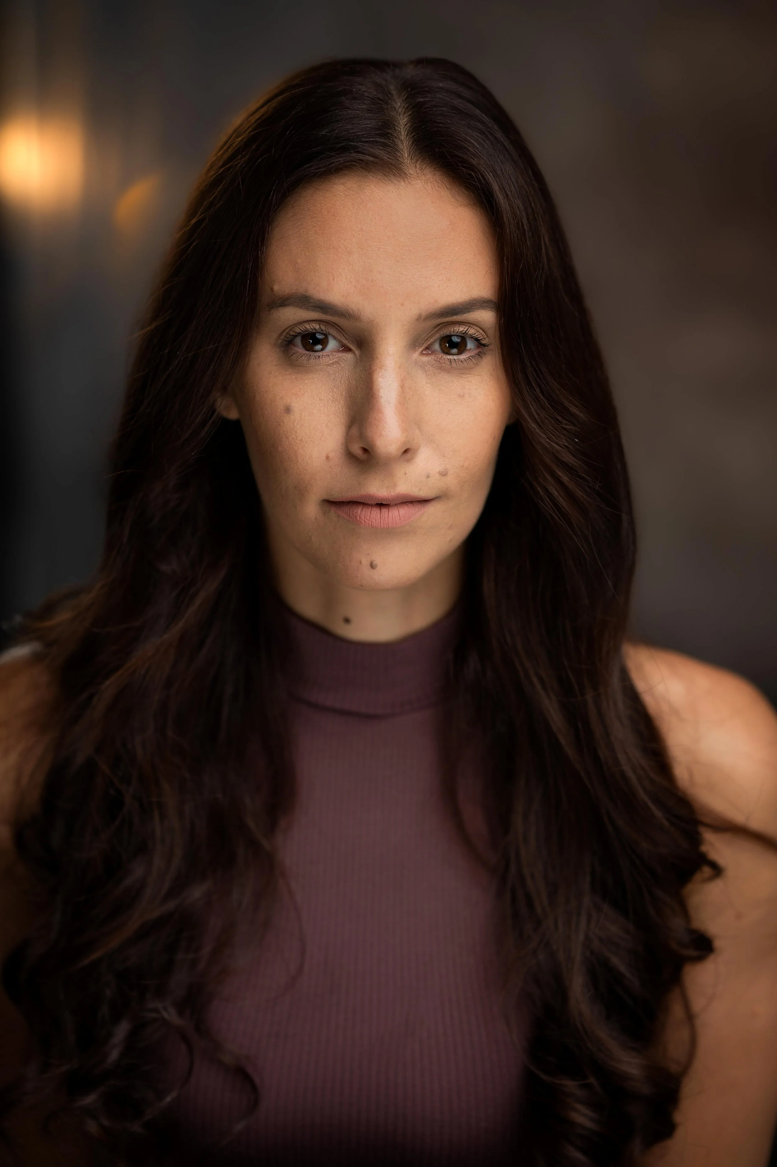 Close-up of a woman with long, dark brown, wavy hair, wearing a sleeveless maroon top, in a softly lit indoor setting.