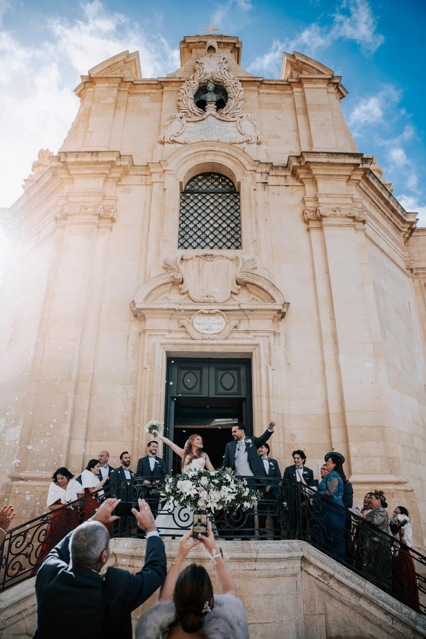 A wedding celebration taking place on the steps of a historic church with ornate architecture, a large bouquet of white flowers, and multiple guests taking photos of the bride and groom who are smiling and waving.