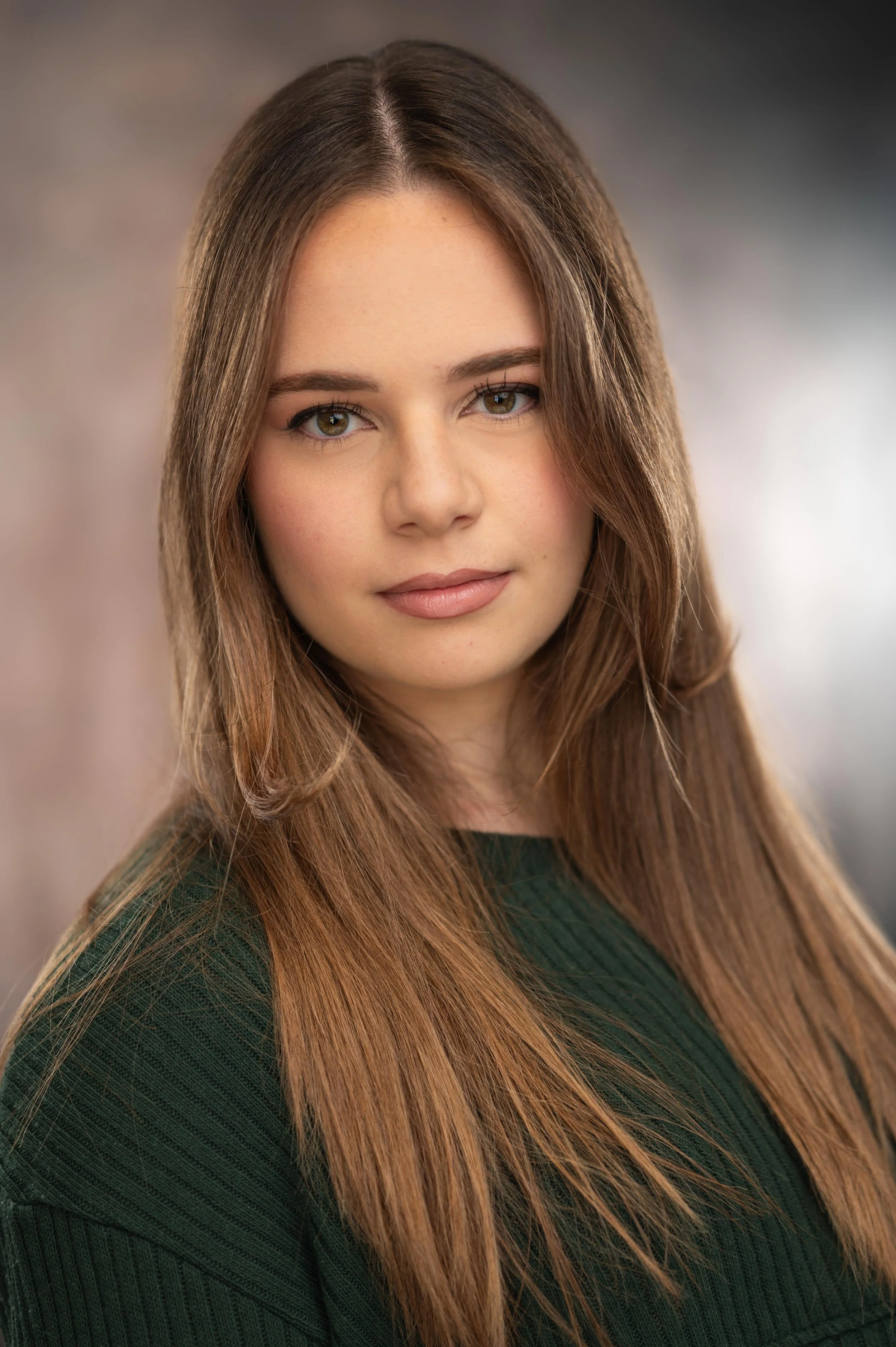 A close-up of a young woman with long, light brown hair, hazel eyes, and fair skin, wearing a dark green ribbed sweater and looking directly at the camera against a neutral background.