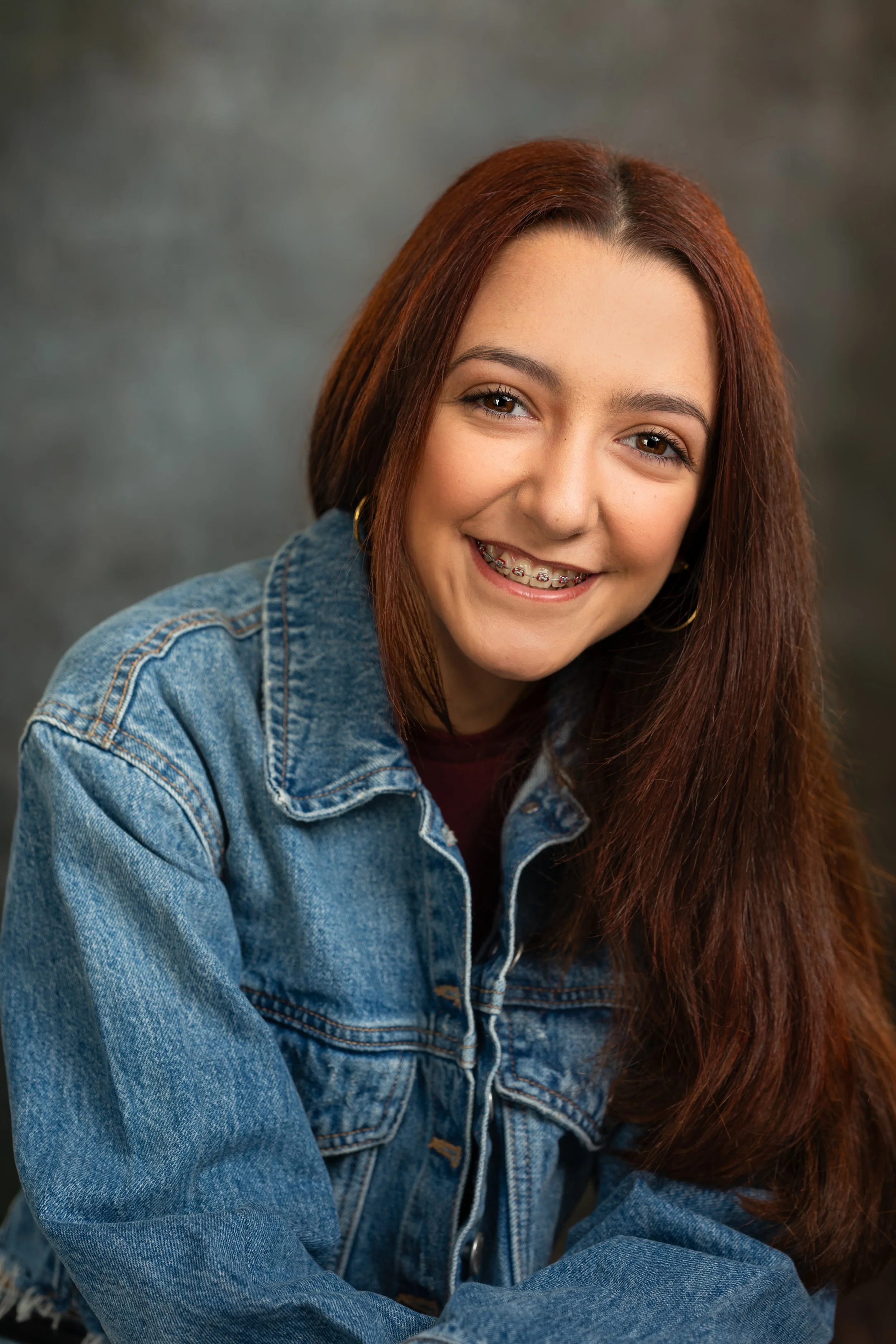 Close-up of a young woman with long red hair, smiling, wearing a denim jacket and braces, against a blurred dark background.
