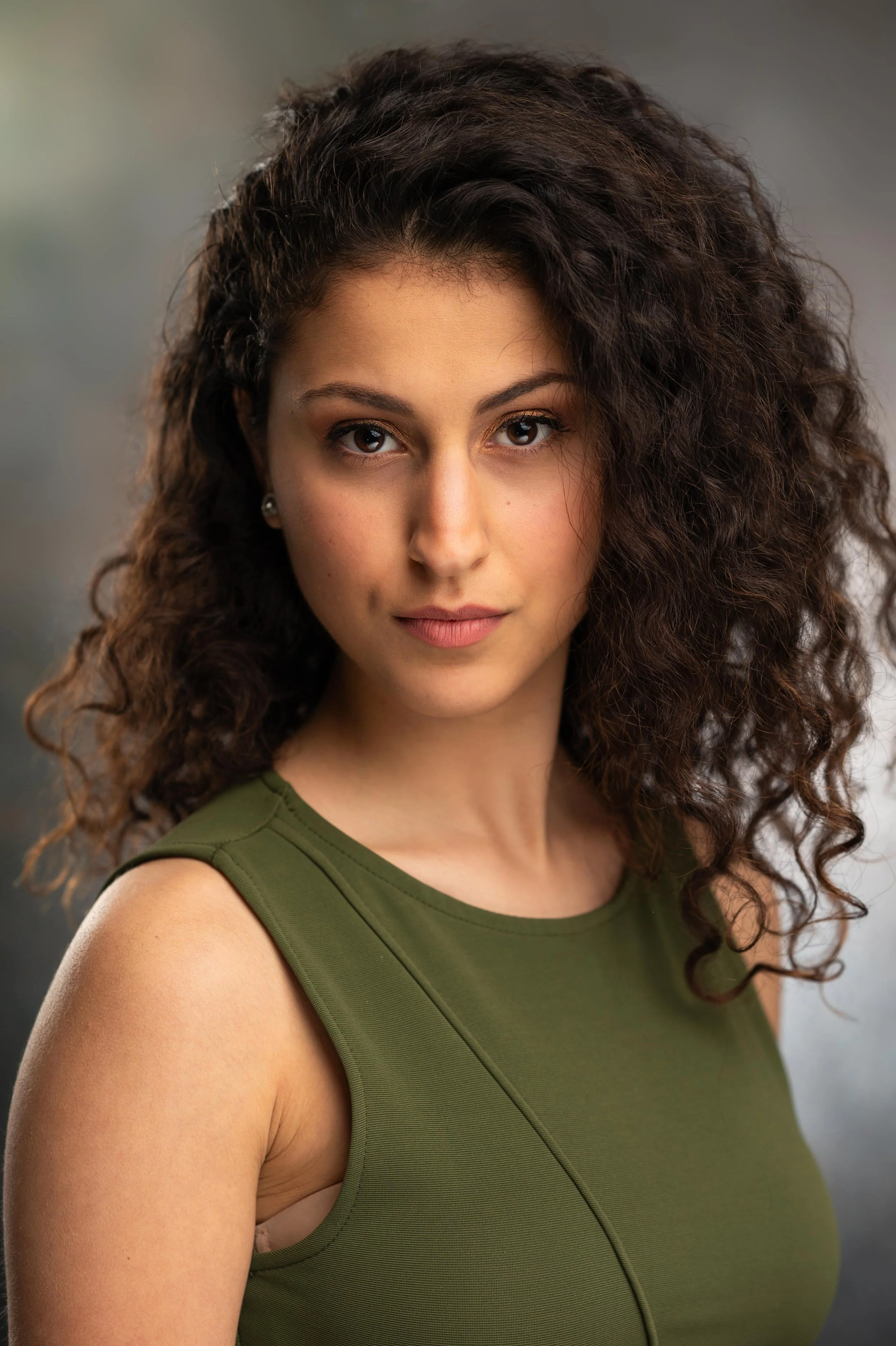 A woman with curly brown hair, wearing a green sleeveless top, looking directly at the camera with a neutral expression.