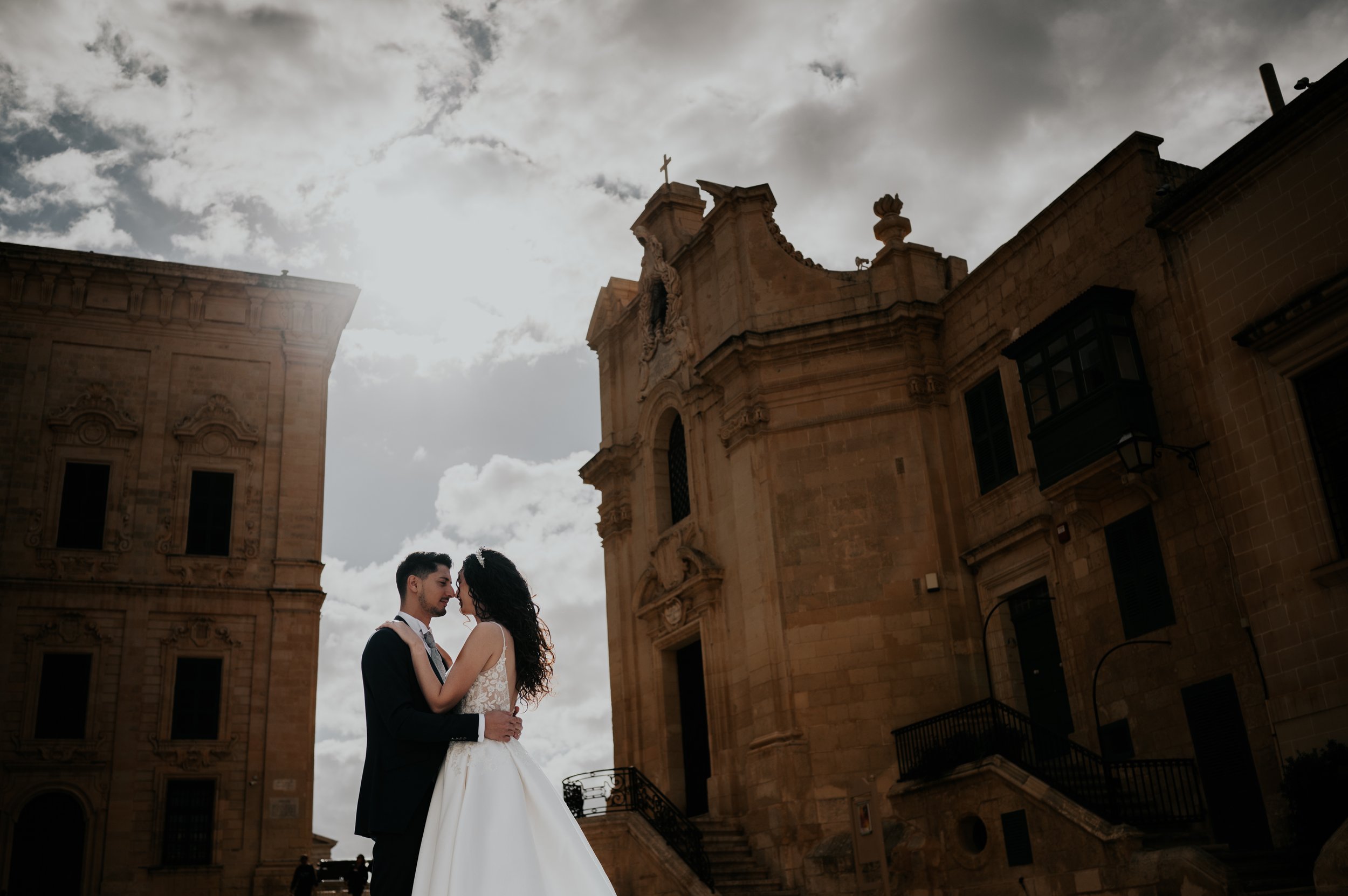A bride and groom are standing close together outside a historic building with a cross on top, cloudy sky in the background.