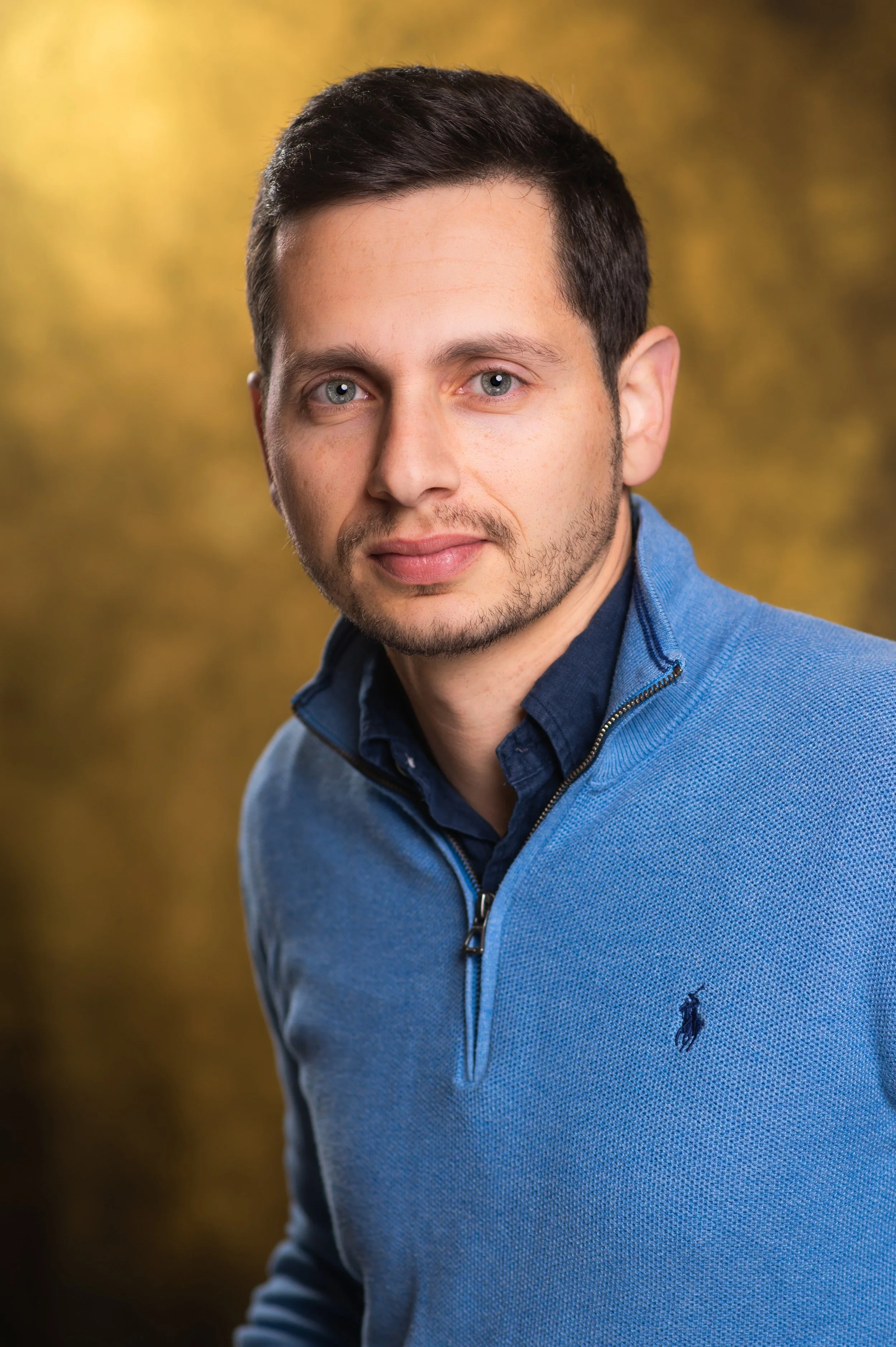 Close-up portrait of a young man with dark hair, blue eyes, and light skin, wearing a blue zip-up sweater with a small logo on the chest, standing outdoors with a blurred autumn background.