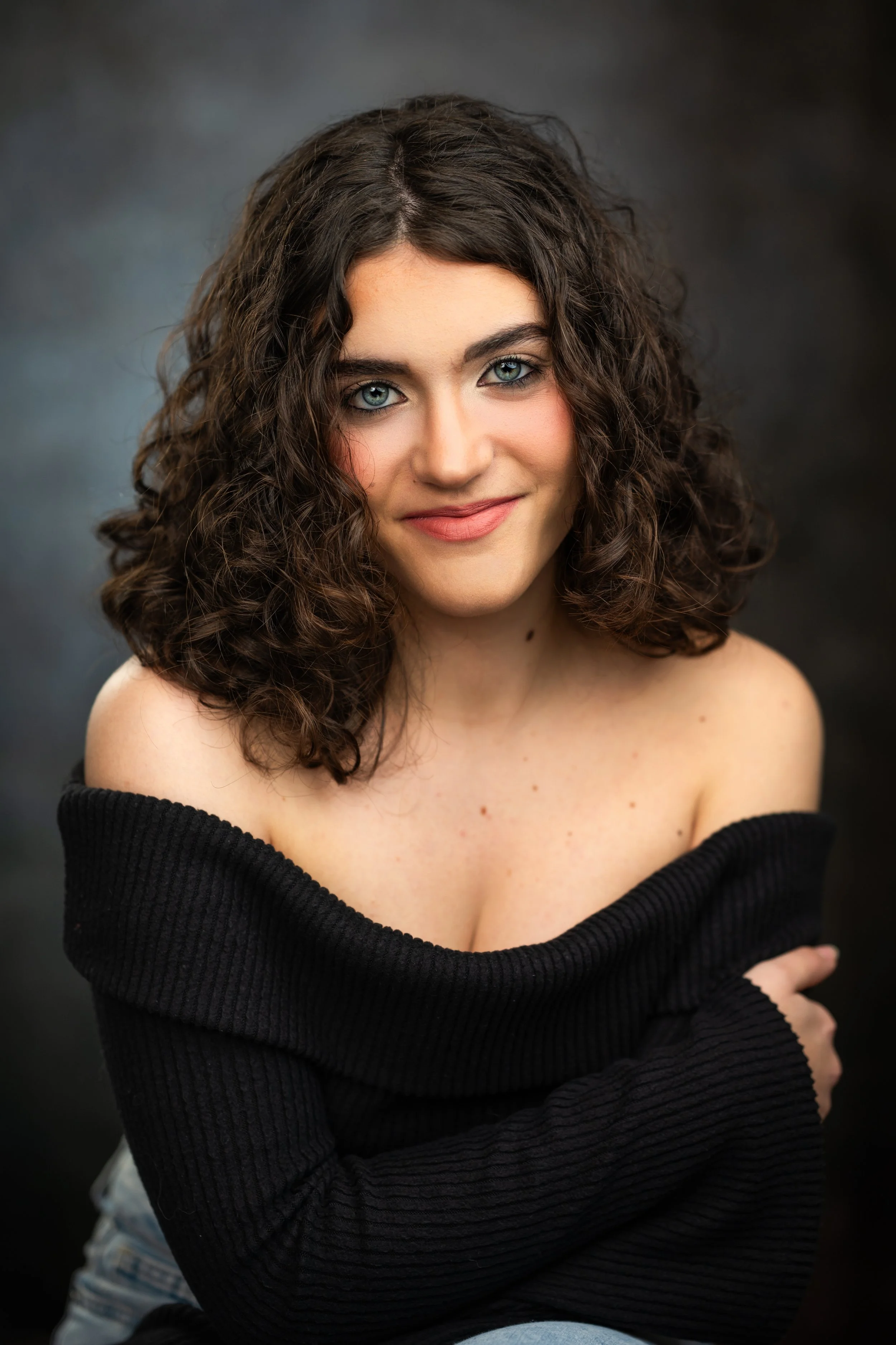 Close-up of a young woman with curly brown hair, blue eyes, light makeup, wearing a black off-the-shoulder sweater, smiling softly against a dark background.