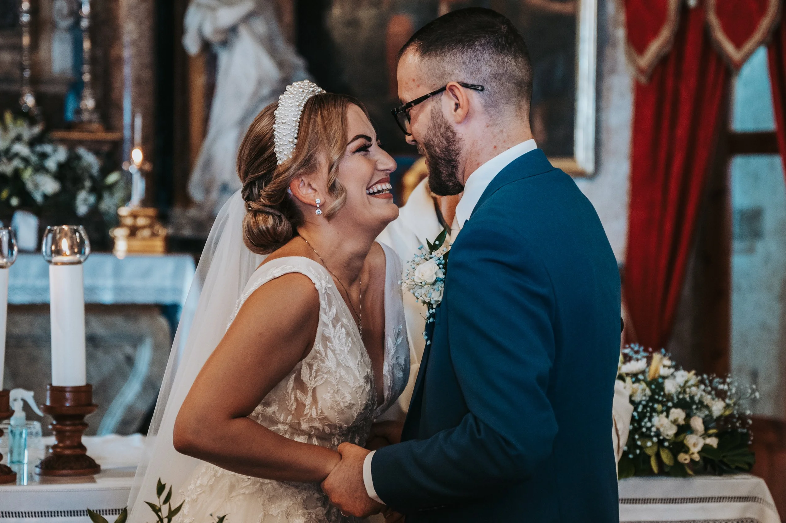 A bride and groom sharing a kiss during their wedding ceremony, holding hands, with wedding decorations and candles in the background.