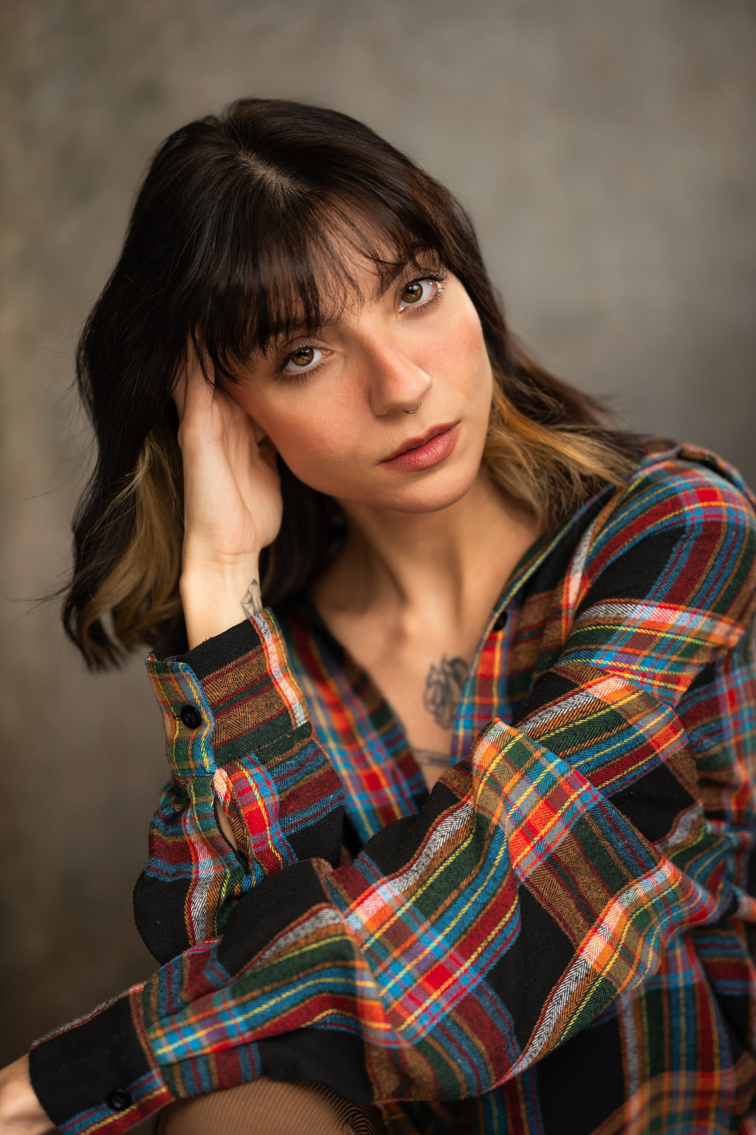 A young woman with brown hair and hazel eyes, wearing a colorful plaid shirt, sitting with her hand resting on her face, looking directly at the camera against a neutral background.