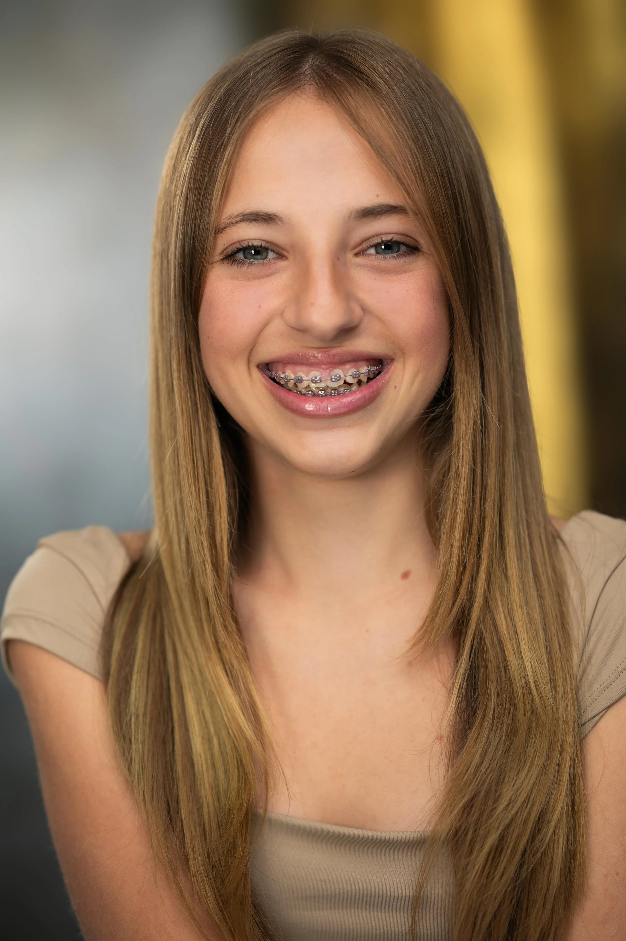 Close-up of a teenage girl smiling, showing braces on her teeth, with long straight hair and wearing a beige top, standing against a blurred indoor background.