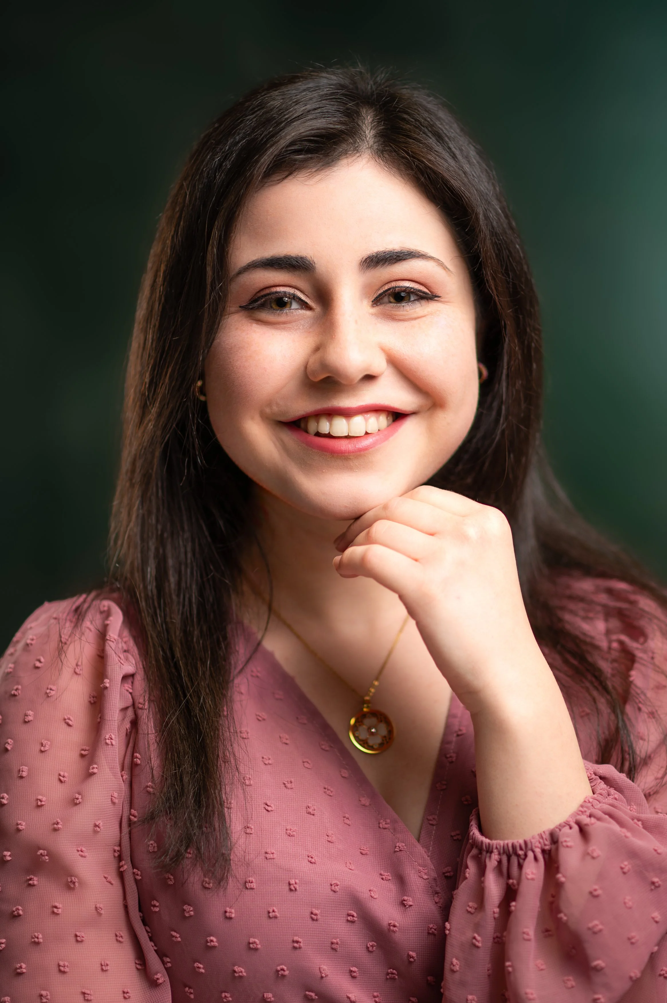 A young woman with long dark hair smiling and resting her hand on her chin, wearing a pink textured blouse and a gold necklace with a round pendant, against a dark green background.