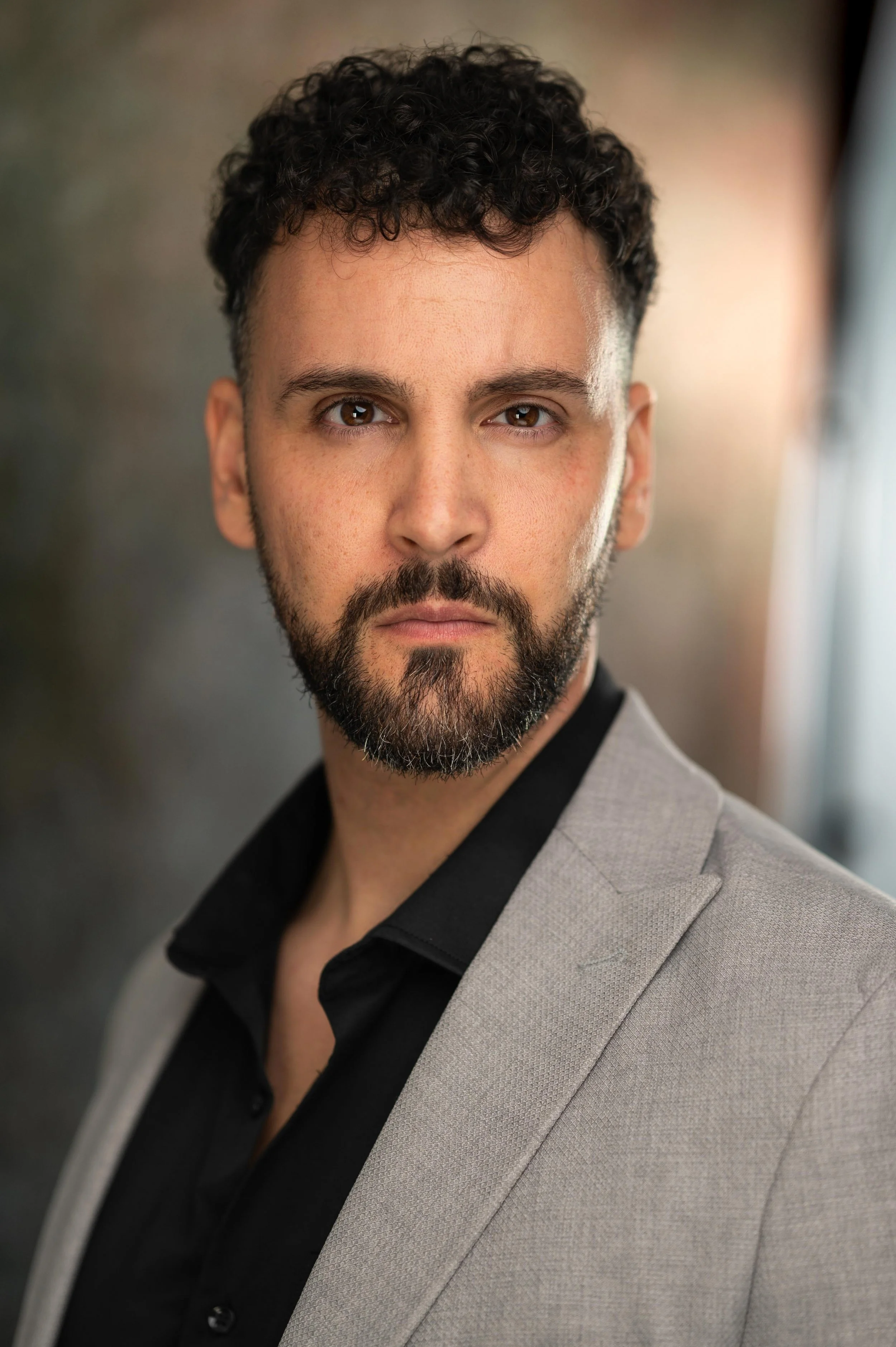 A man with curly dark hair and a beard wearing a light gray suit jacket and a black shirt, looking directly at the camera with a serious expression.