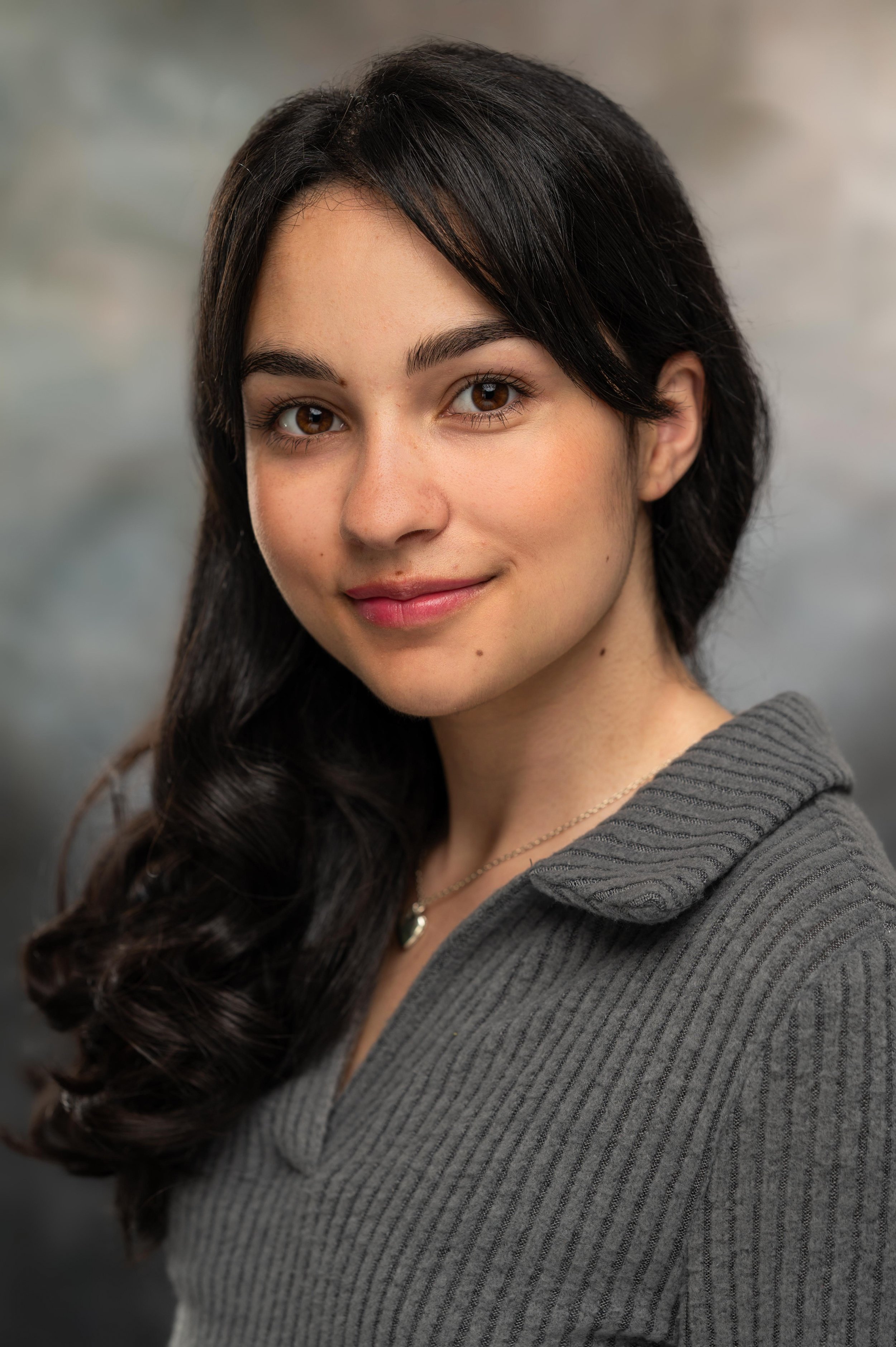 Close-up of a young woman with long, wavy dark hair, brown eyes, and a subtle smile, wearing a gray ribbed sweater and a gold necklace, against a blurred neutral background.