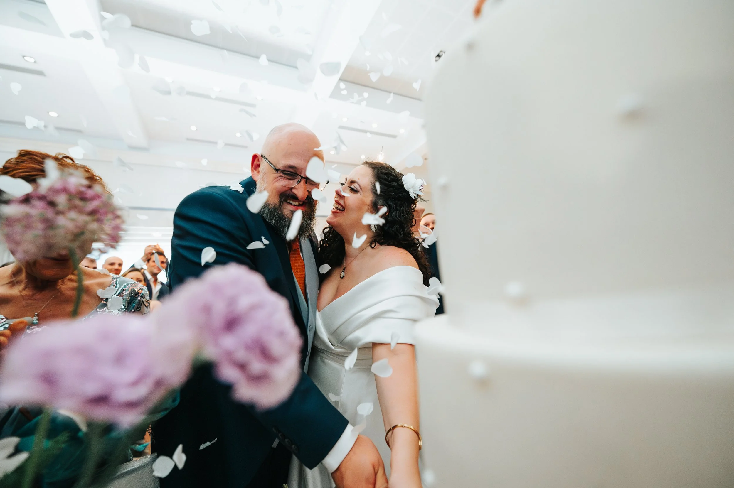 A bride and groom smiling and dancing at their wedding reception, surrounded by falling white confetti and guests.