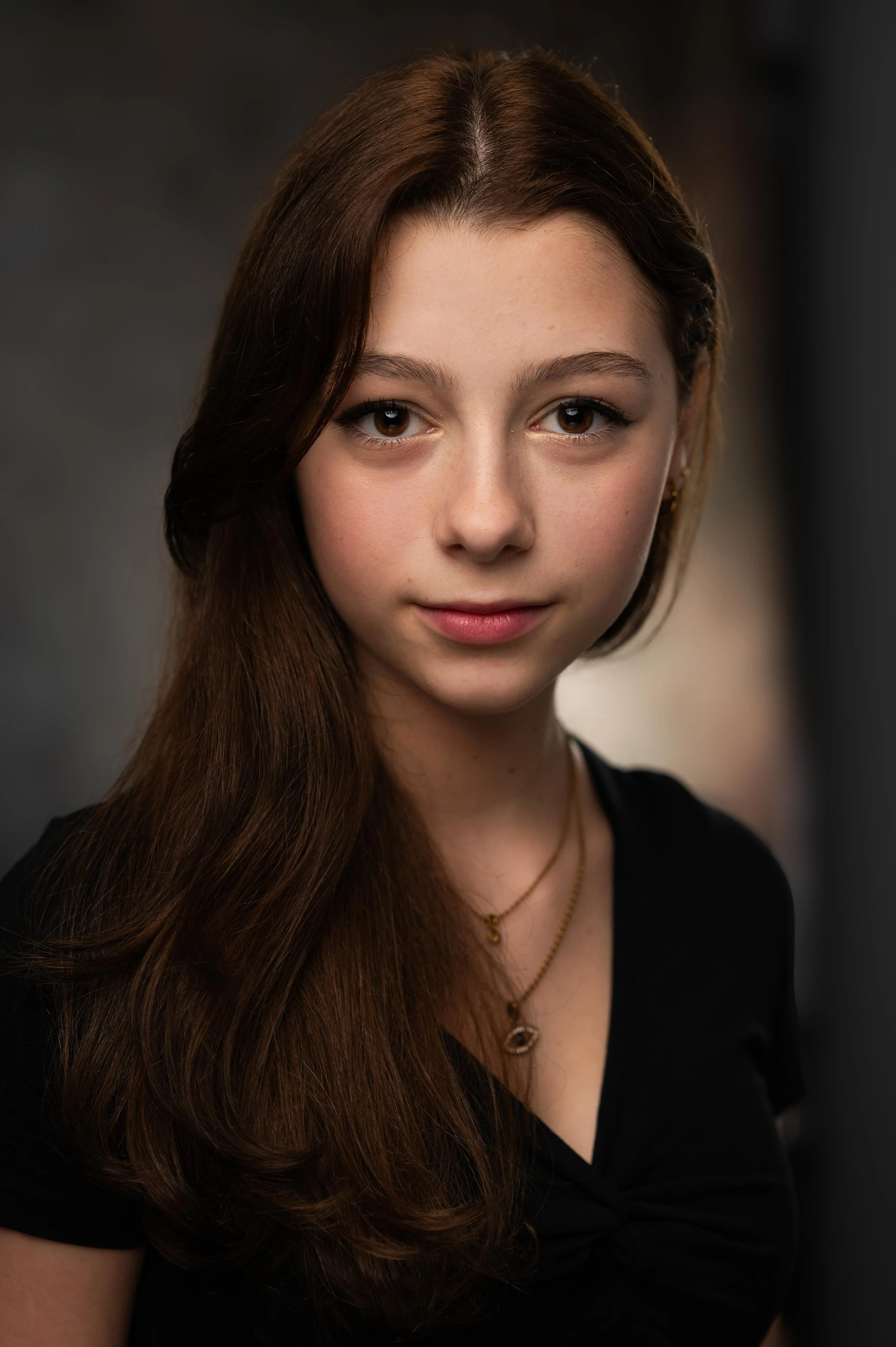 Close-up portrait of a young woman with long brown hair, wearing a black top and layered gold necklaces, looking into the camera with a slight smile.