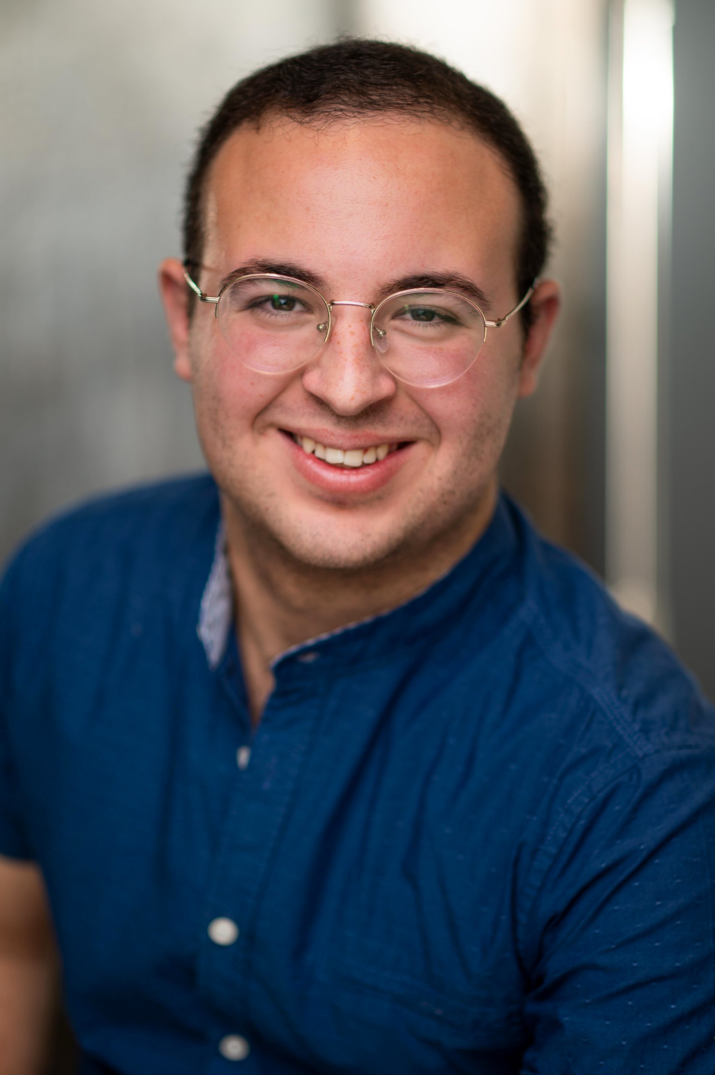 Close-up of a young man with short dark hair, wearing round eyeglasses and a blue button-up shirt, smiling at the camera.