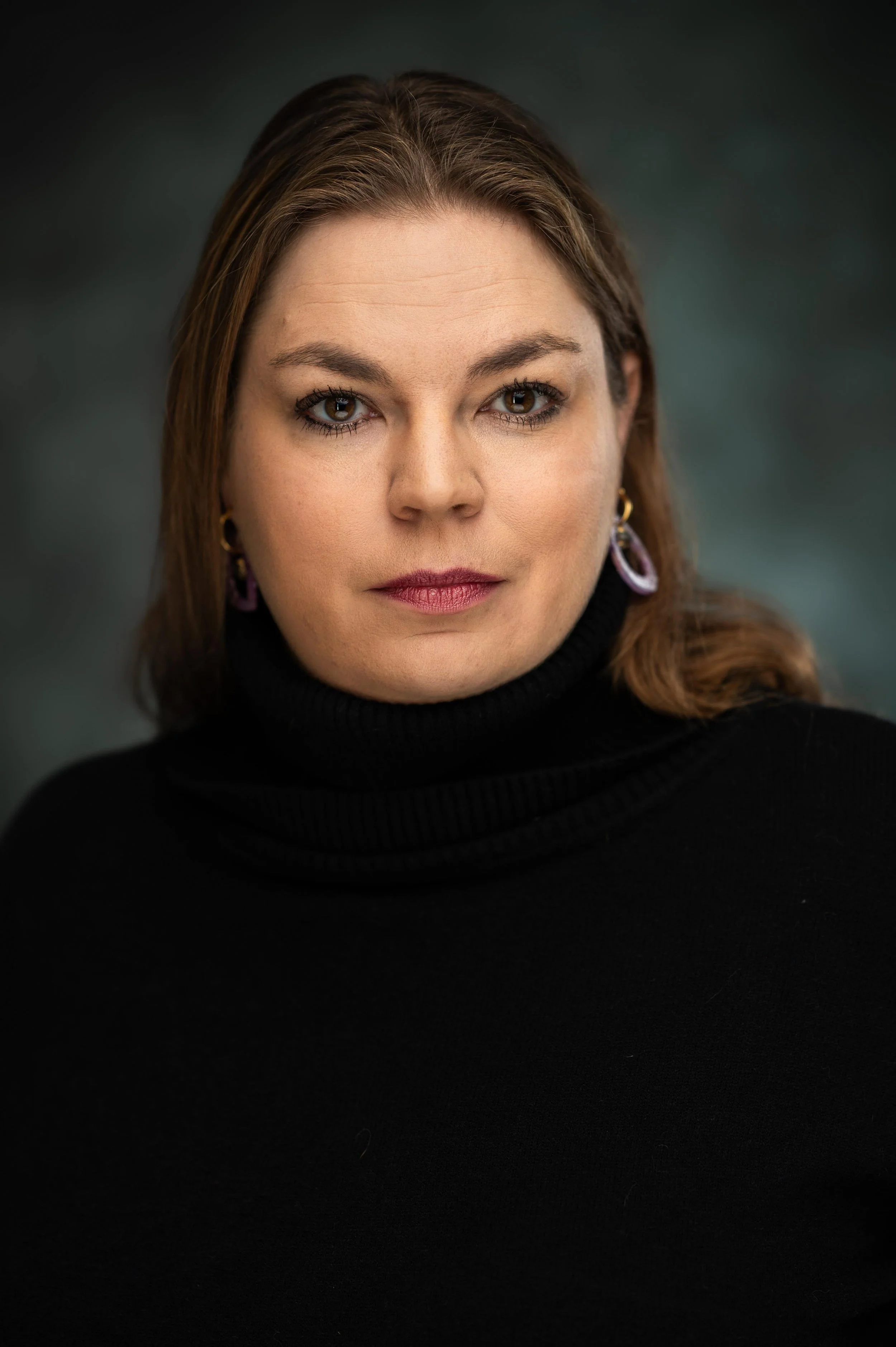 Close-up portrait of a woman with brown hair, brown eyes, wearing a black turtleneck sweater and purple earrings, looking directly at the camera.