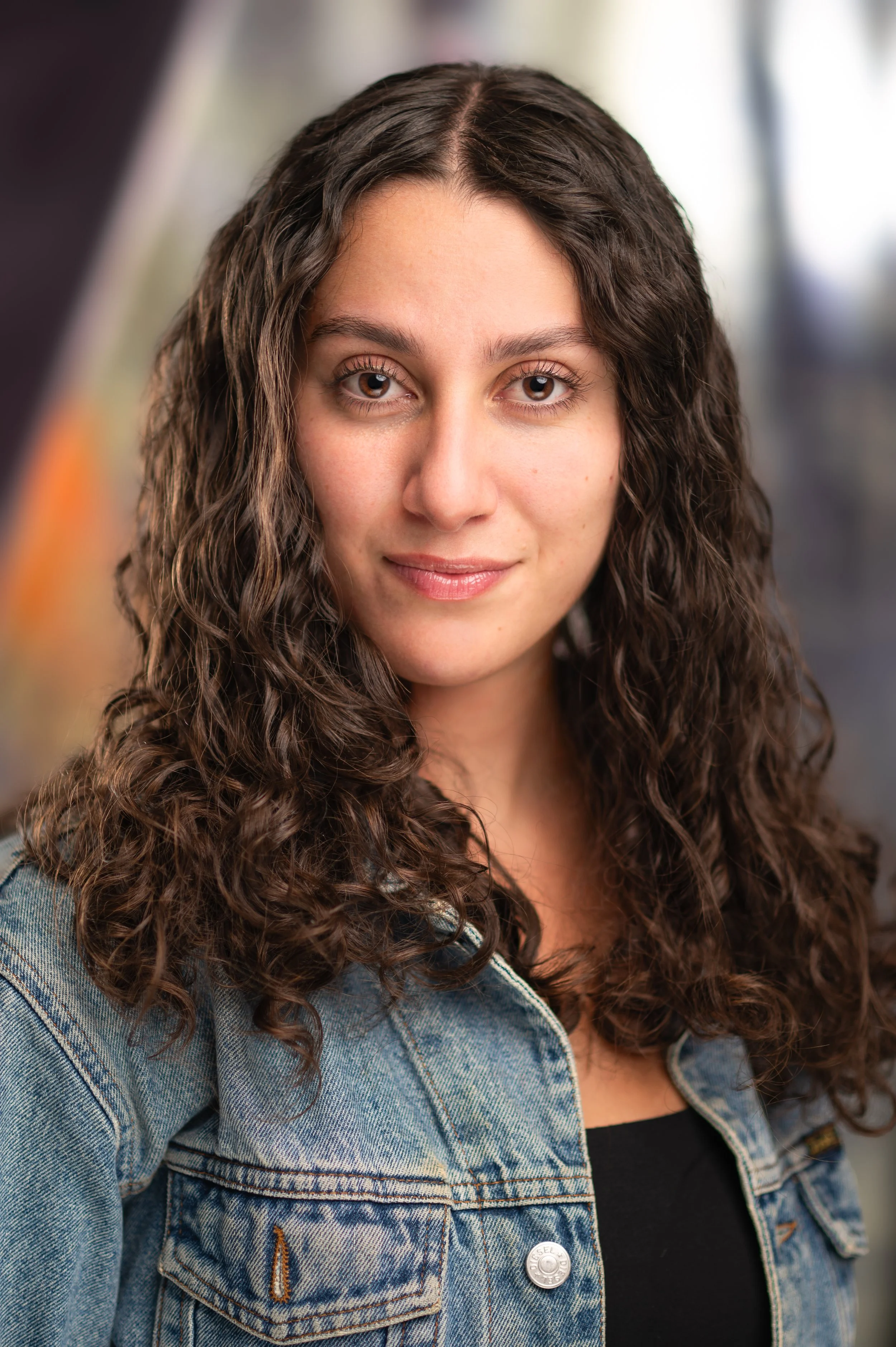 A young woman with long curly brown hair, wearing a denim jacket over a black top, looking directly into the camera with a slight smile.