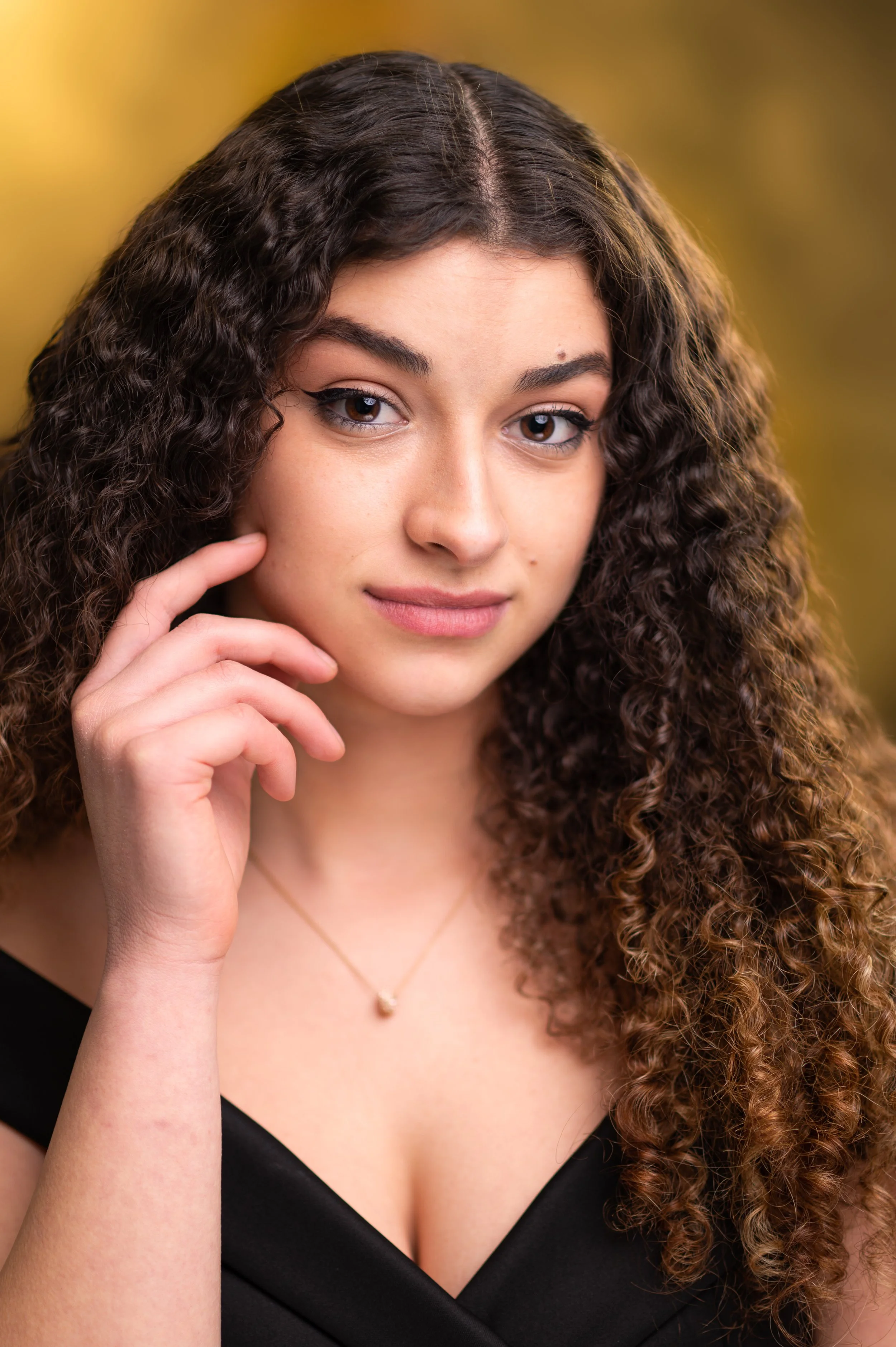 Close-up of a young woman with curly brown hair, touching her face near her jawline, wearing a black top and a delicate gold necklace with a small pendant, against a blurred yellowish background.