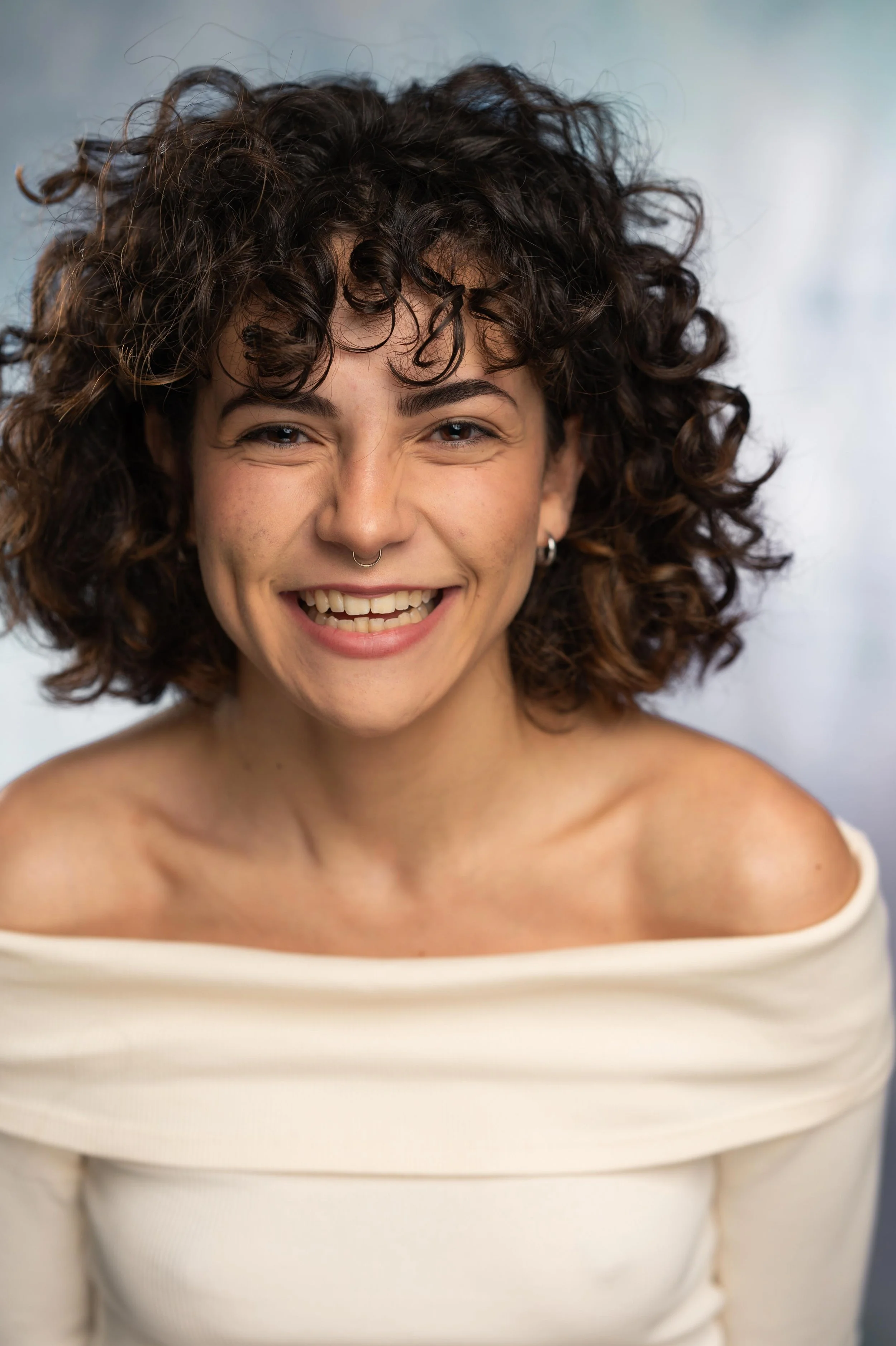 A woman with curly dark hair, smiling widely, wearing a white off-the-shoulder top, and showing her teeth.