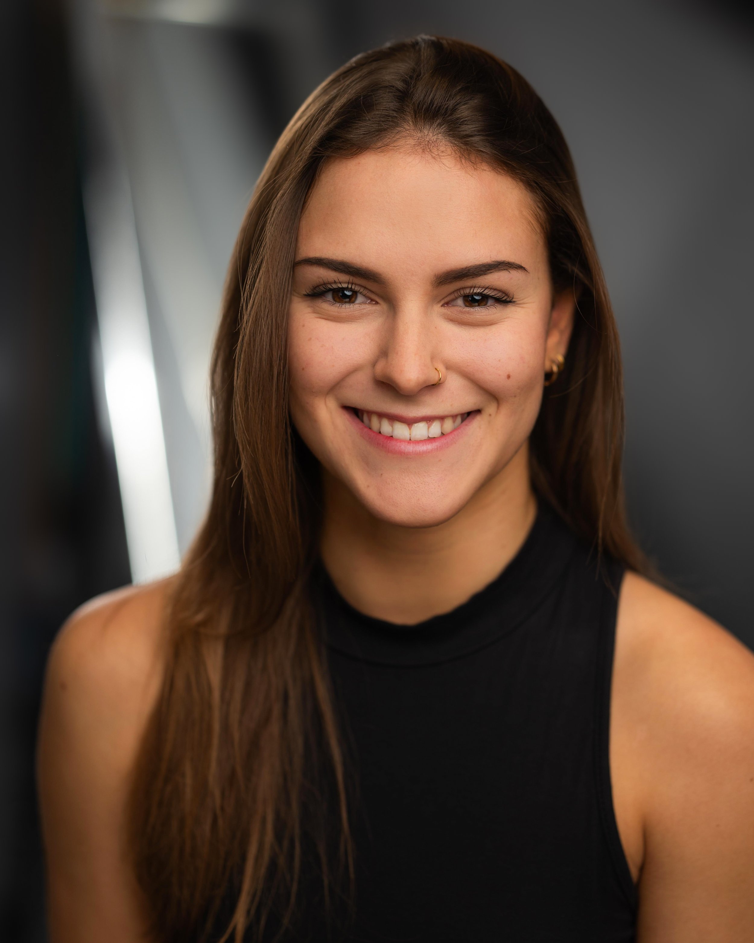 Close-up of a young woman with long brown hair, smiling, wearing a black sleeveless top, with a blurred background.