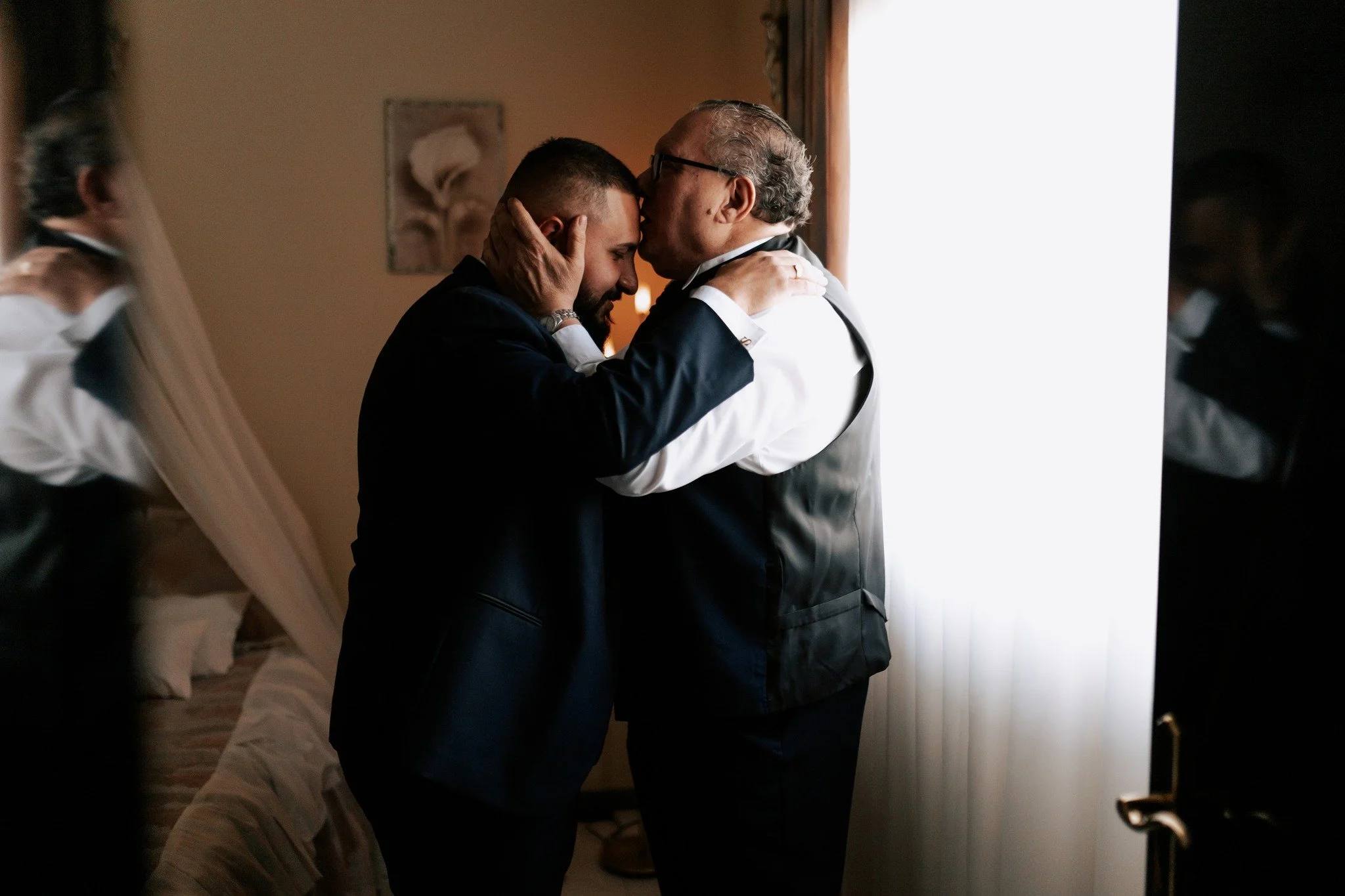 An older man and a younger man, both dressed in formal attire including bow ties, are sharing an emotional moment by holding each other's heads with their faces close together, likely in a bedroom with a bed and a painting visible in the background.
