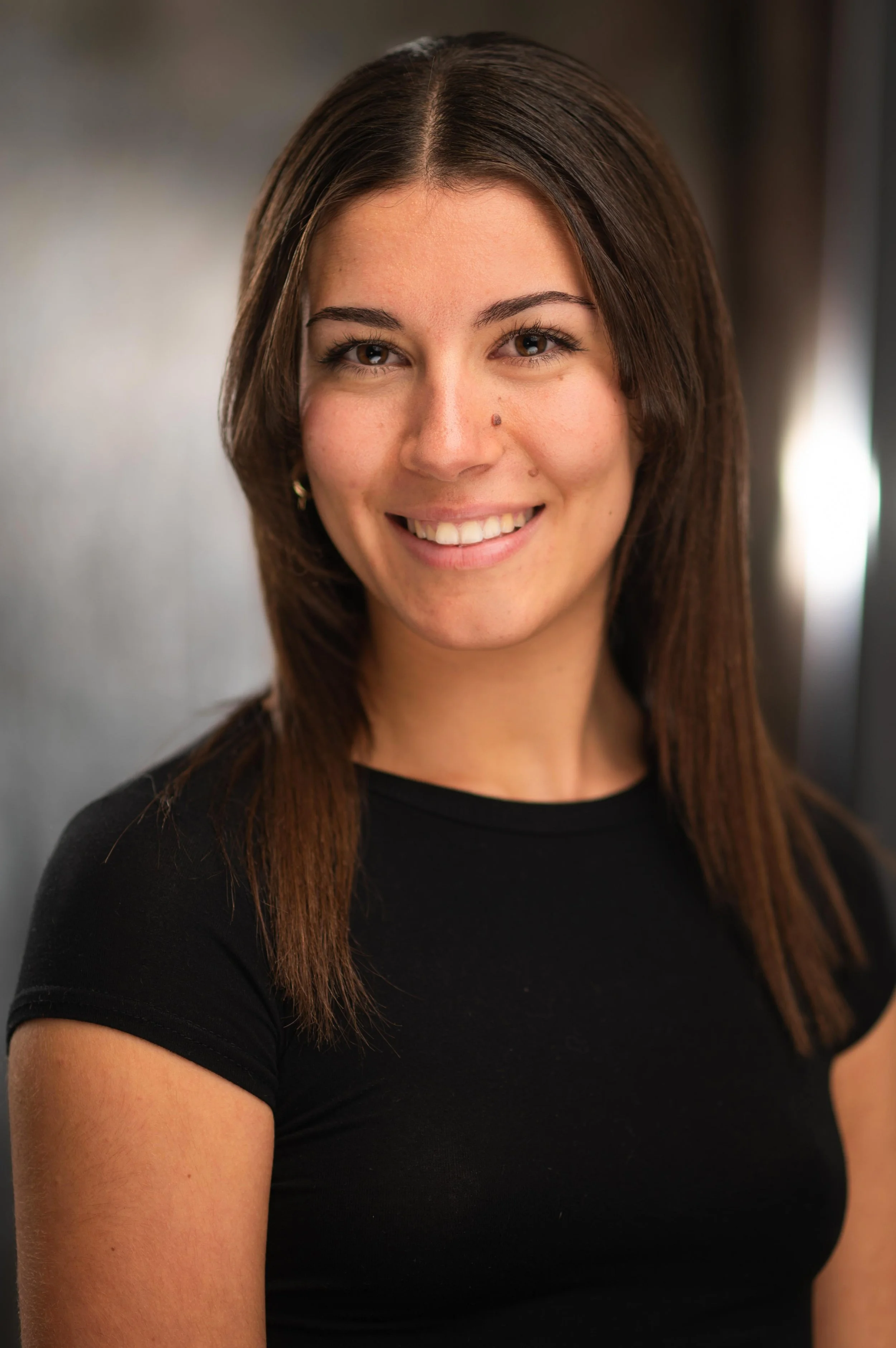 Close-up portrait of a woman with long brown hair, brown eyes, and a nose piercing, smiling and wearing a black top.