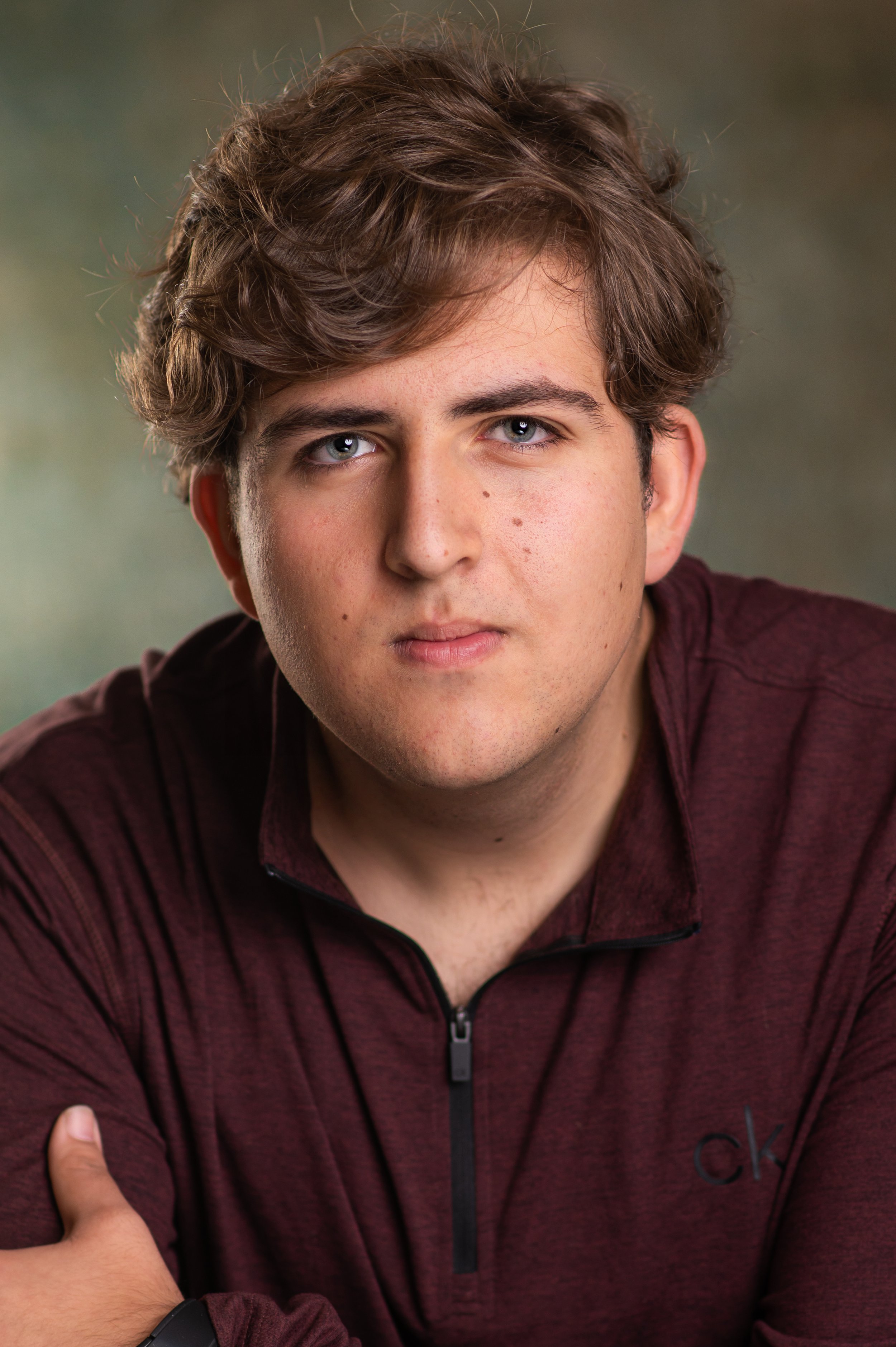 Young man with curly brown hair and blue eyes wearing a maroon zip-up jacket, facing forward with arms crossed, against a blurred green background.