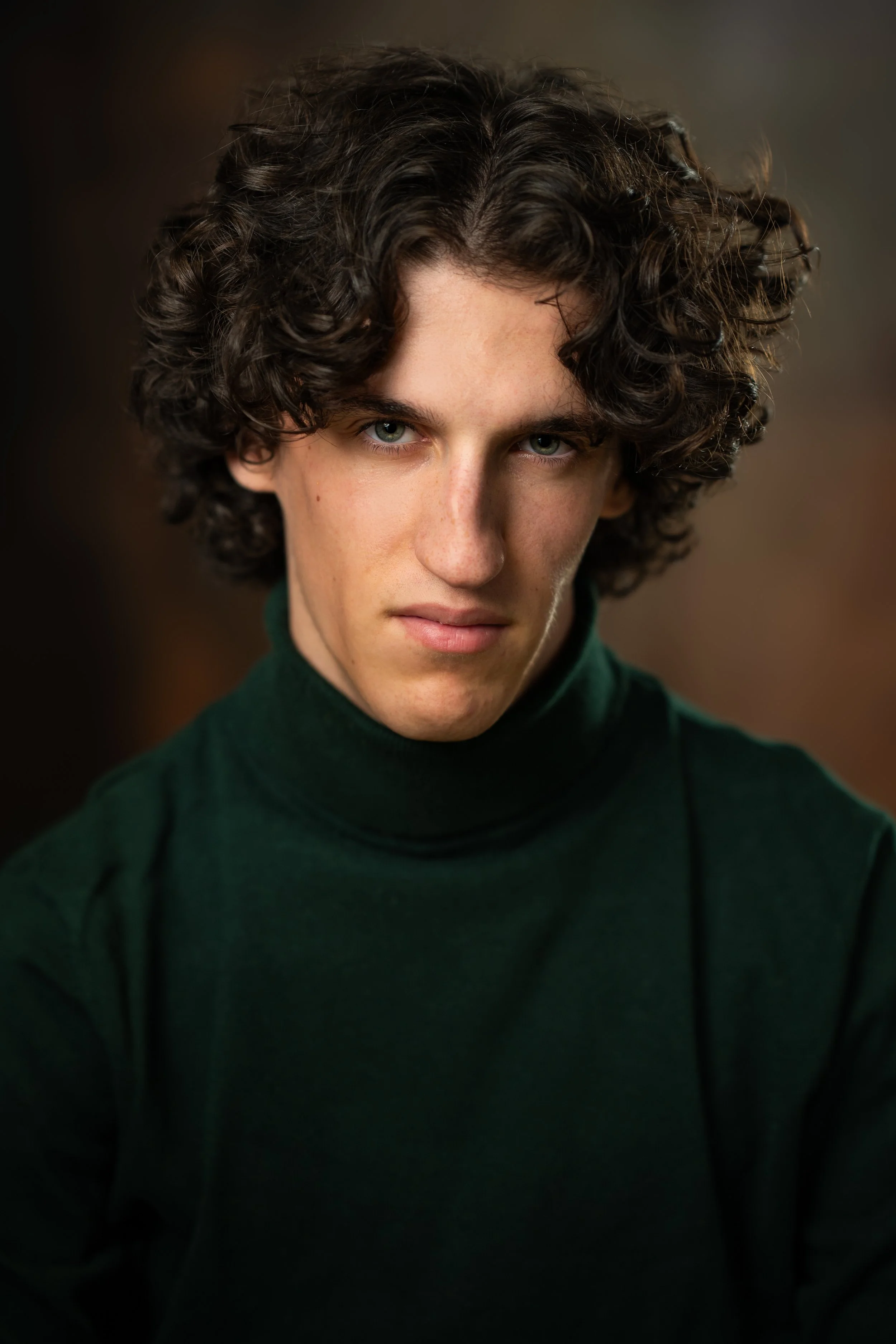 Close-up of a young man with curly dark hair and piercing blue eyes, wearing a dark green turtleneck sweater, looking directly at the camera against a blurred background.