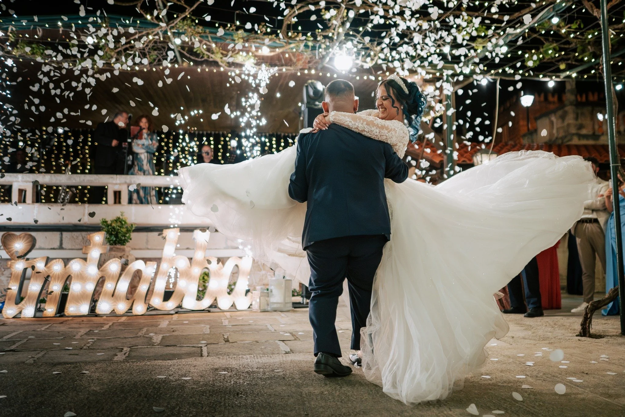 A newlywed couple dancing at their wedding reception, with the bride in a white wedding dress and the groom in a dark tuxedo. Fairy lights and decorations are visible in the background, and there are people watching in the distance.