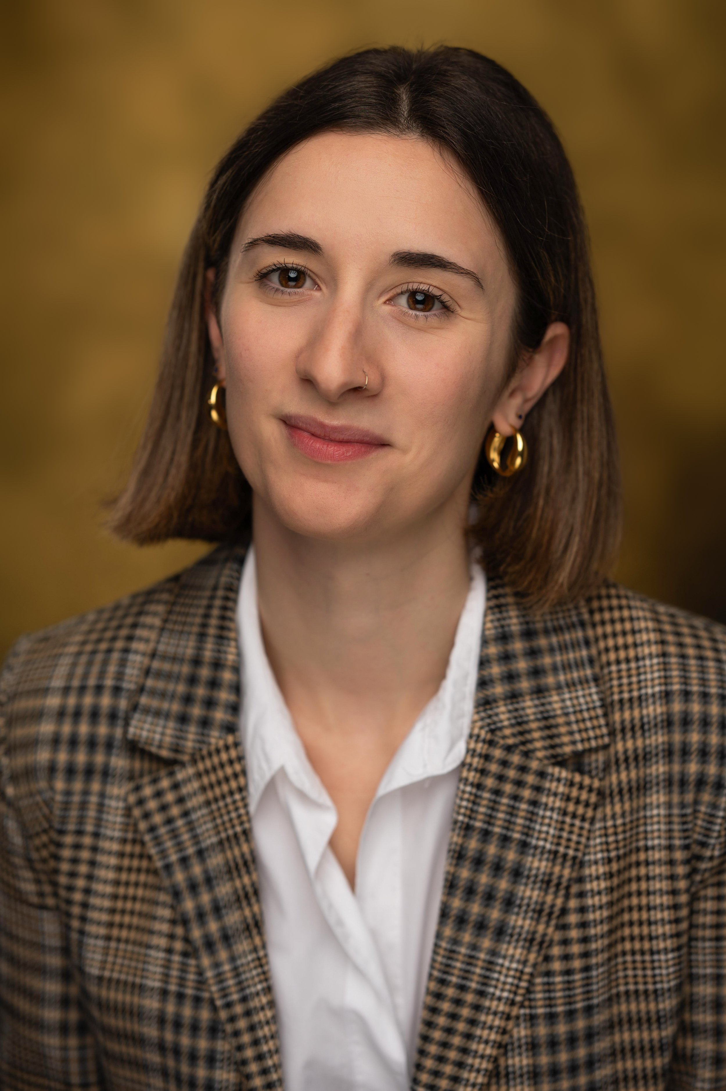 A woman with brown hair, wearing gold hoop earrings, a white shirt, and a checkered blazer, smiling softly with a blurred autumn-colored background.
