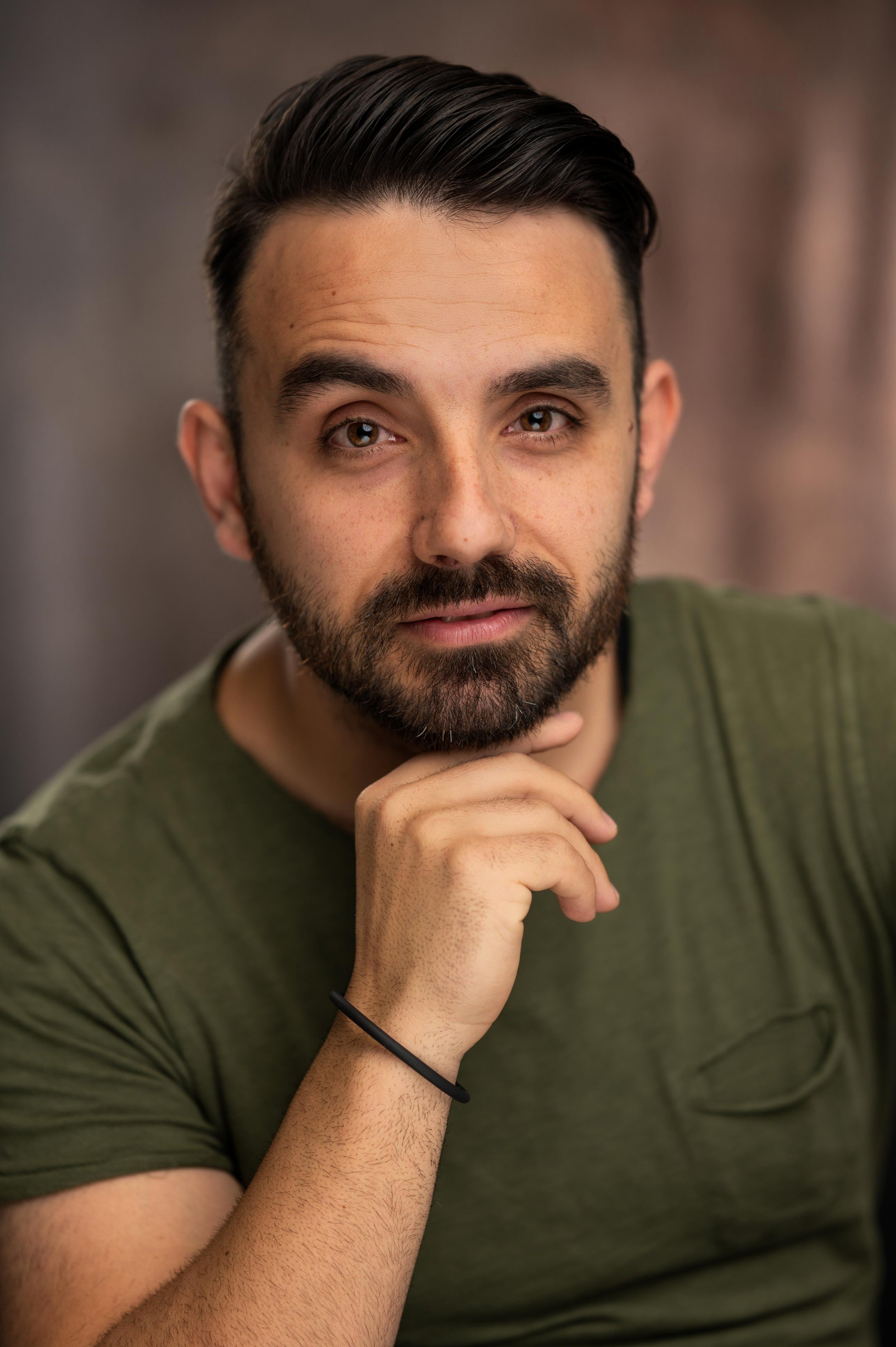 A man with dark hair and a beard, wearing an olive green shirt, resting his chin on his hand, with a neutral background.
