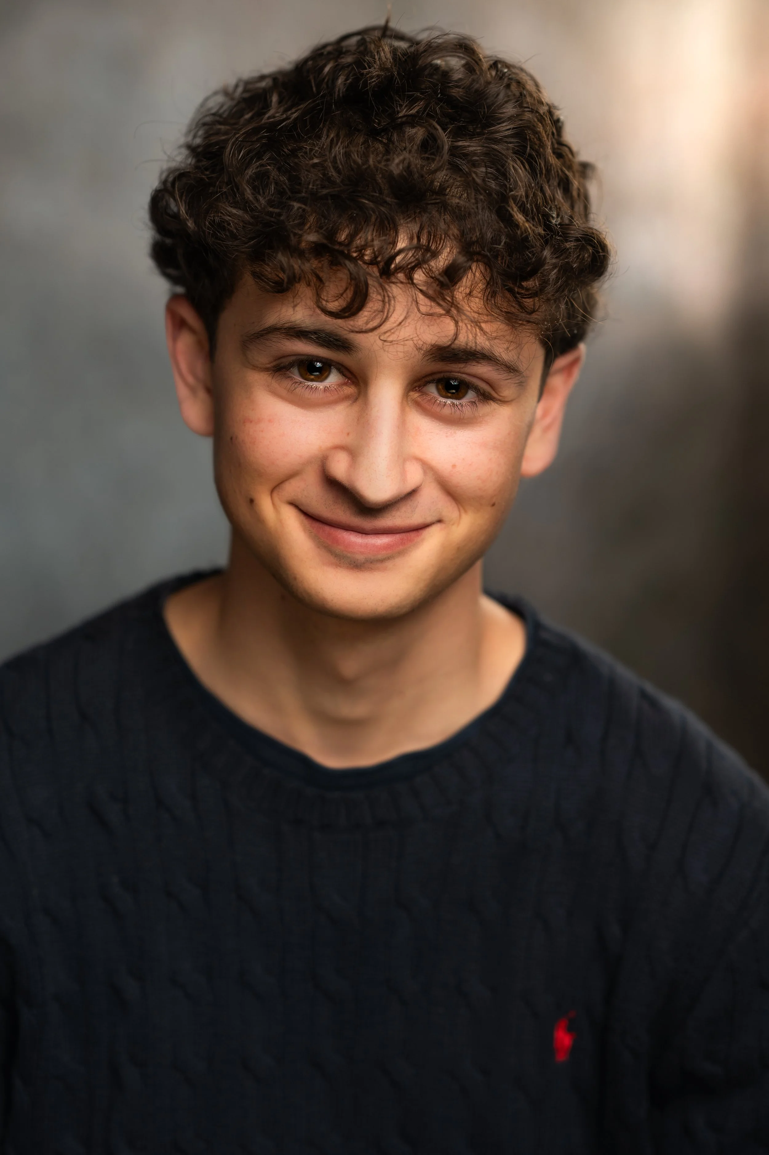 Close-up of a young man with curly brown hair and brown eyes, smiling gently, wearing a dark sweater with a small red logo on the chest, against a blurred gray background.