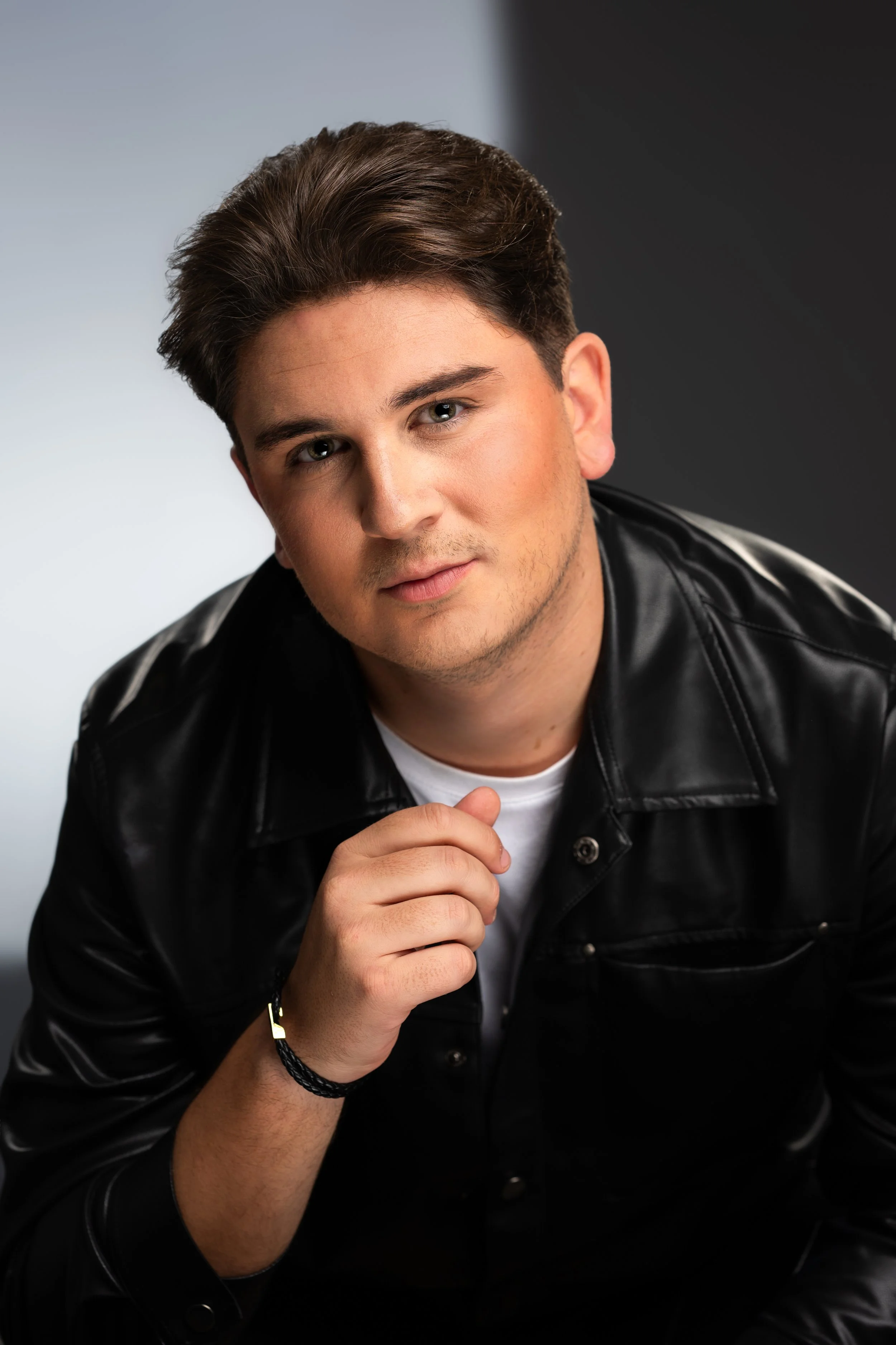 Portrait of a young man with short dark hair wearing a black leather jacket and white T-shirt, looking directly at the camera with a neutral expression, against a background featuring a split of light gray and black.