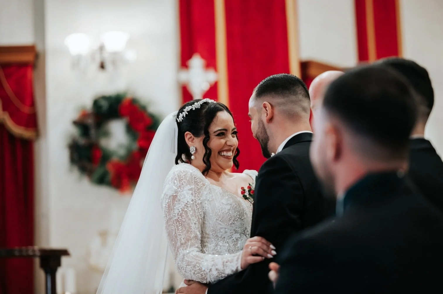 A bride and groom holding hands and smiling during their wedding ceremony indoors, with red curtains and holiday decorations in the background.