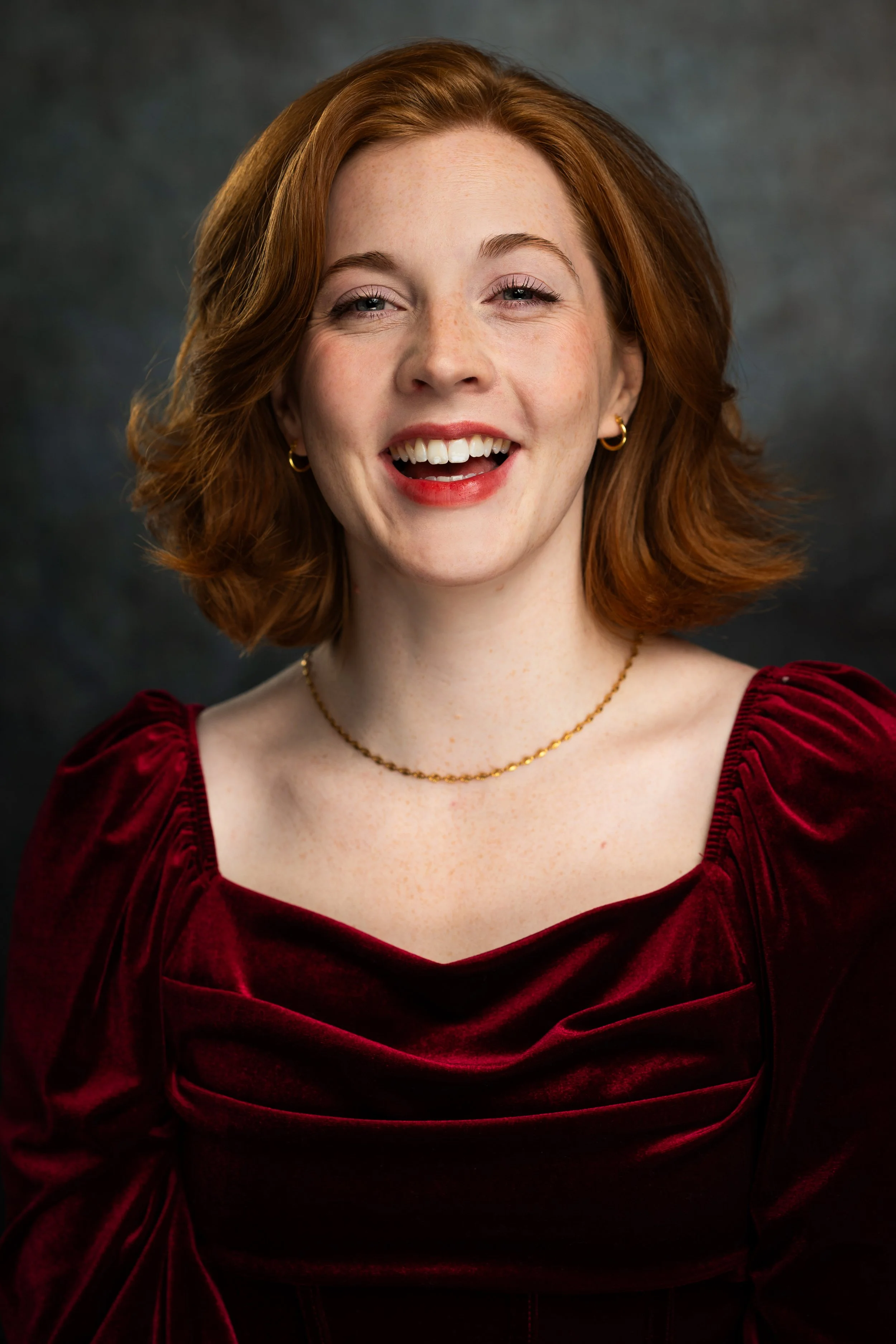 Close-up of a smiling woman with red hair, wearing a red velvet dress, gold hoop earrings, and a delicate gold necklace, against a dark background.