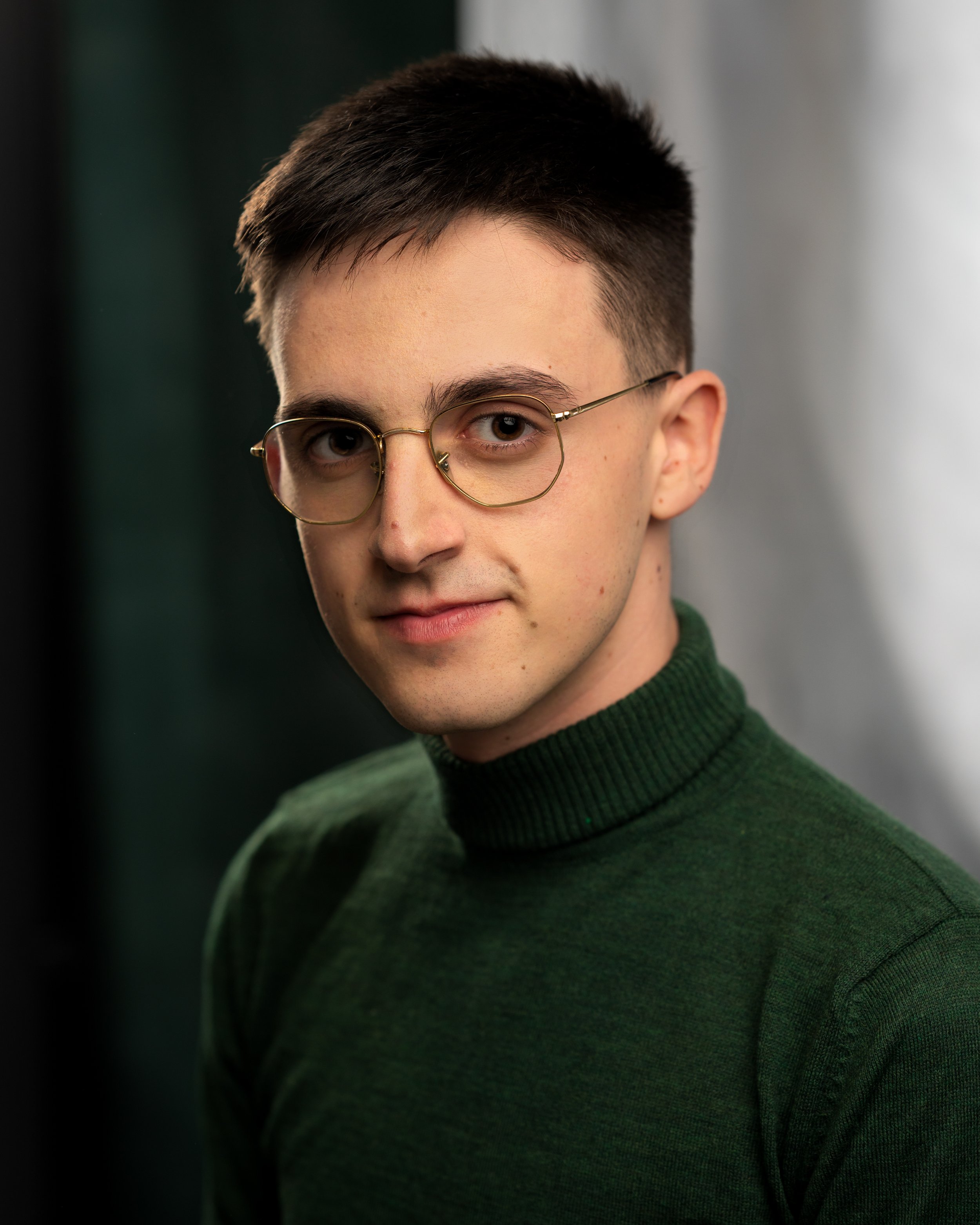 Close-up of a young man with short dark hair, wearing round gold-rimmed glasses and a dark green sweater, looking at the camera with a slight smile, indoor setting with blurred background.