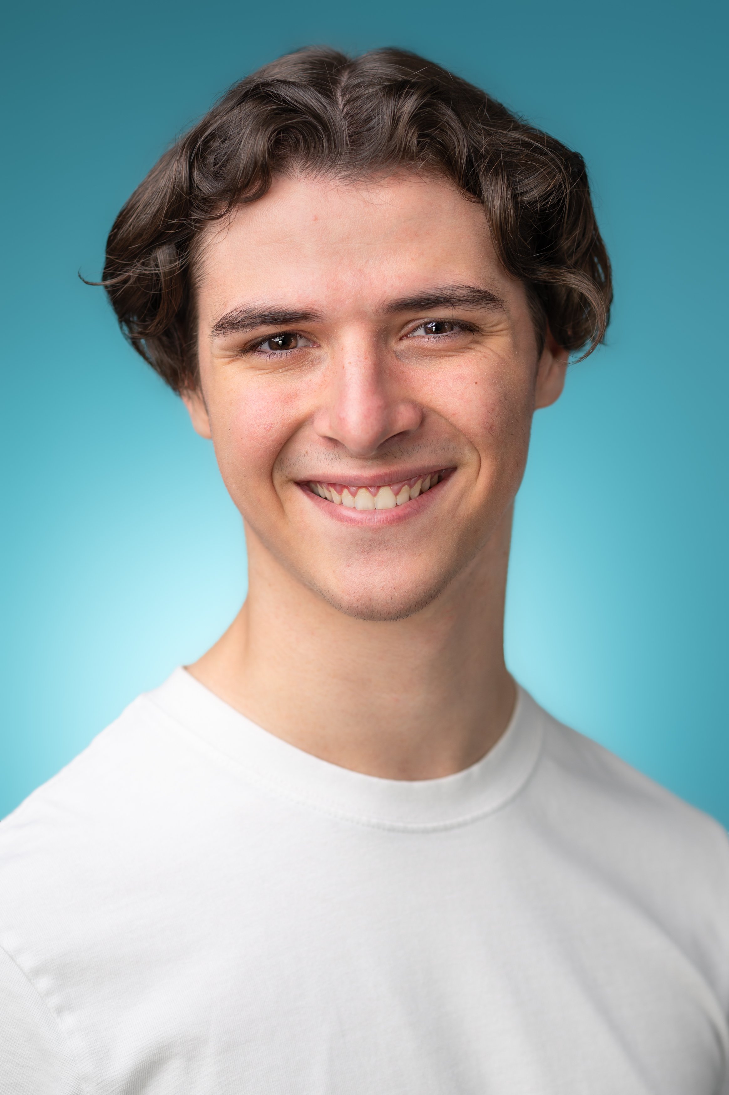 Close-up portrait of a smiling young man with brown curly hair, wearing a white t-shirt, against a blue background.
