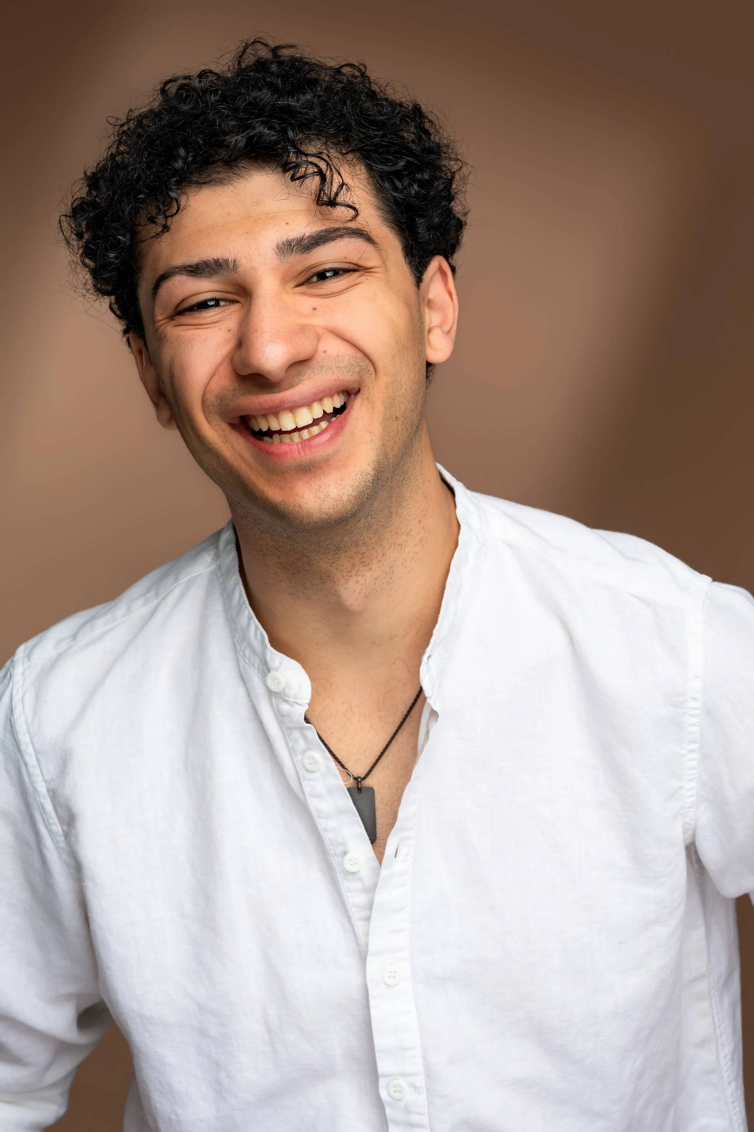 A young man with curly hair smiling, wearing a white button-up shirt and a black necklace with a rectangular pendant, against a plain, brown background.