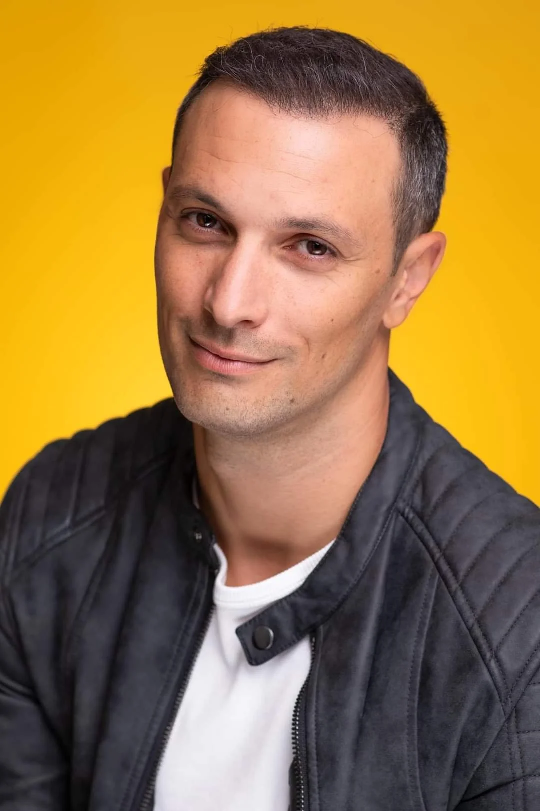 Portrait of a man with short dark hair, light skin, in a black leather jacket and white shirt, smiling slightly against a yellow background.