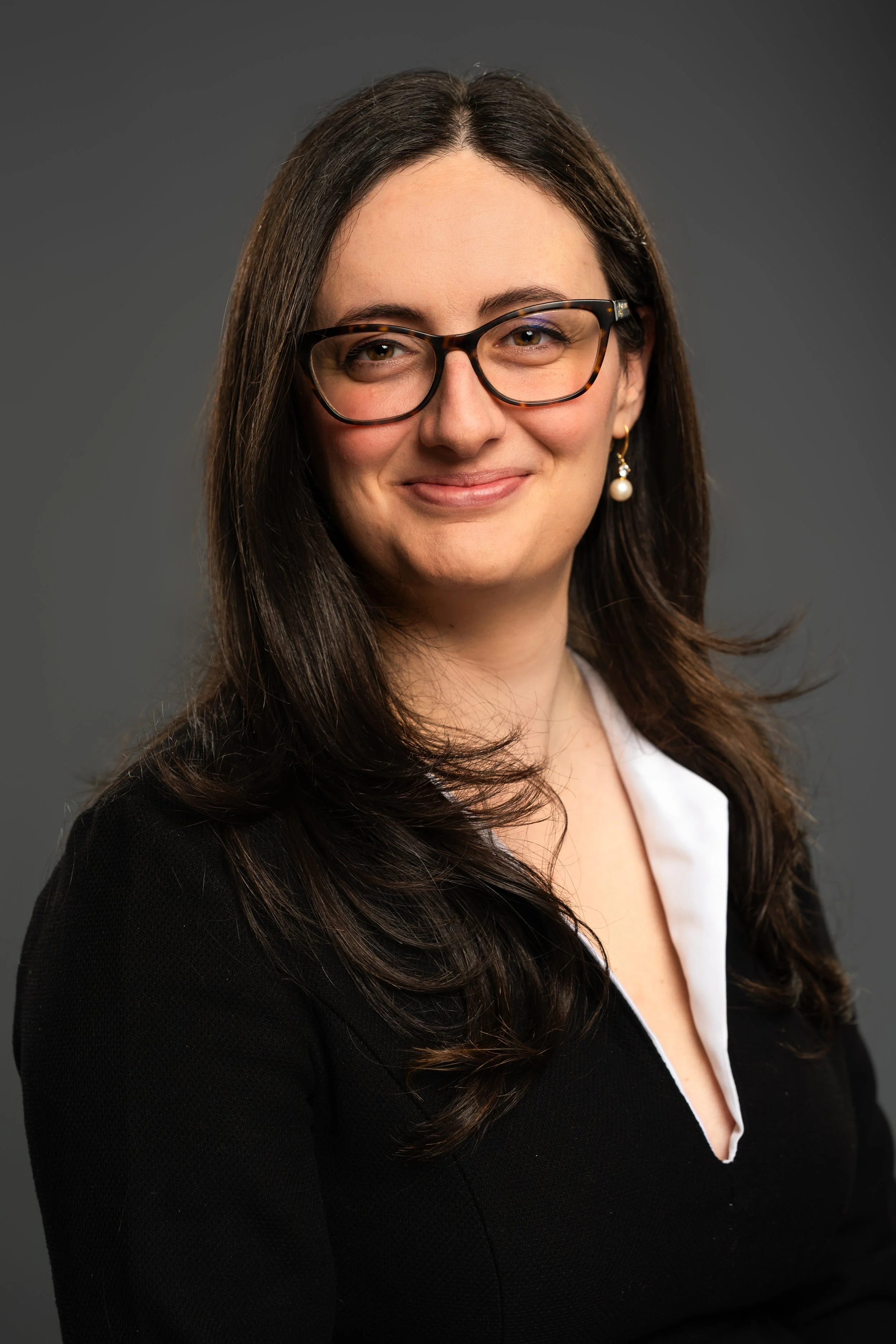 A professional woman with long brown hair, wearing glasses, pearl earrings, a white shirt, and a black blazer, smiling against a dark gray background.