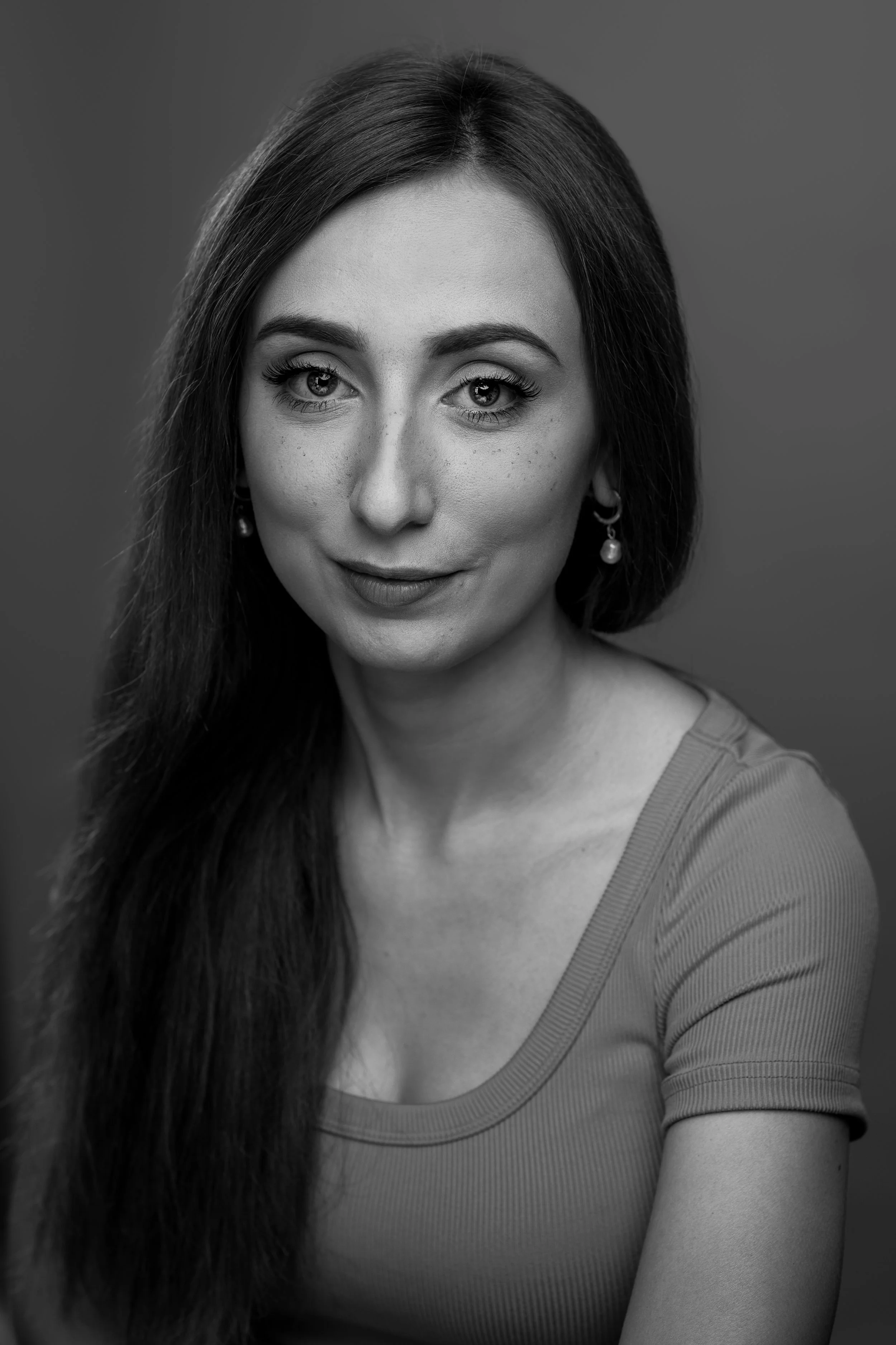 A black and white portrait of a woman with long dark hair, wearing a top with a rounded neckline, and earrings. She is looking directly at the camera with a slight smile.