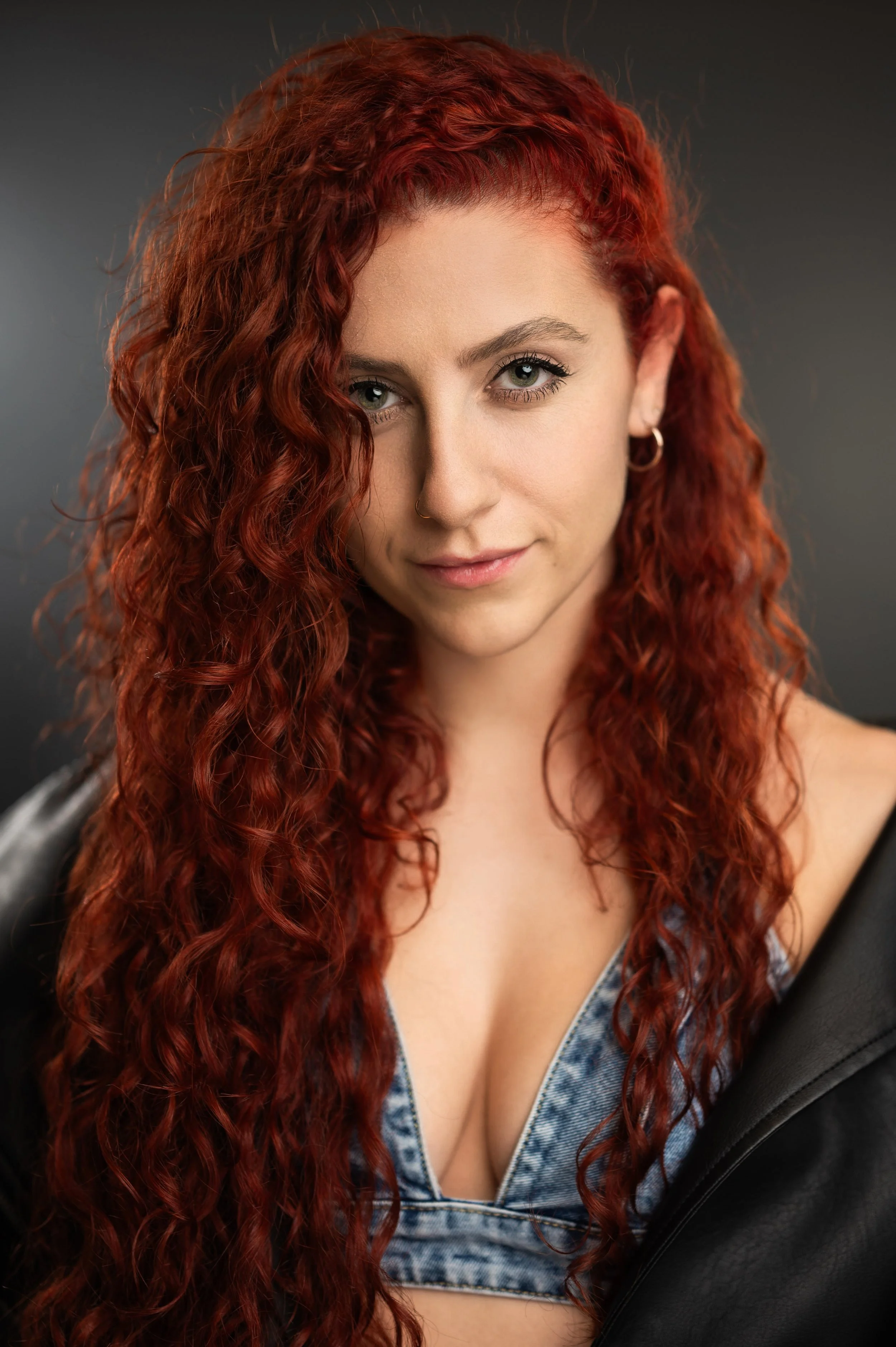 Close-up portrait of a woman with long, curly red hair, green eyes, wearing hoop earrings, a nose ring, a denim top, and a black leather jacket, against a gray background.